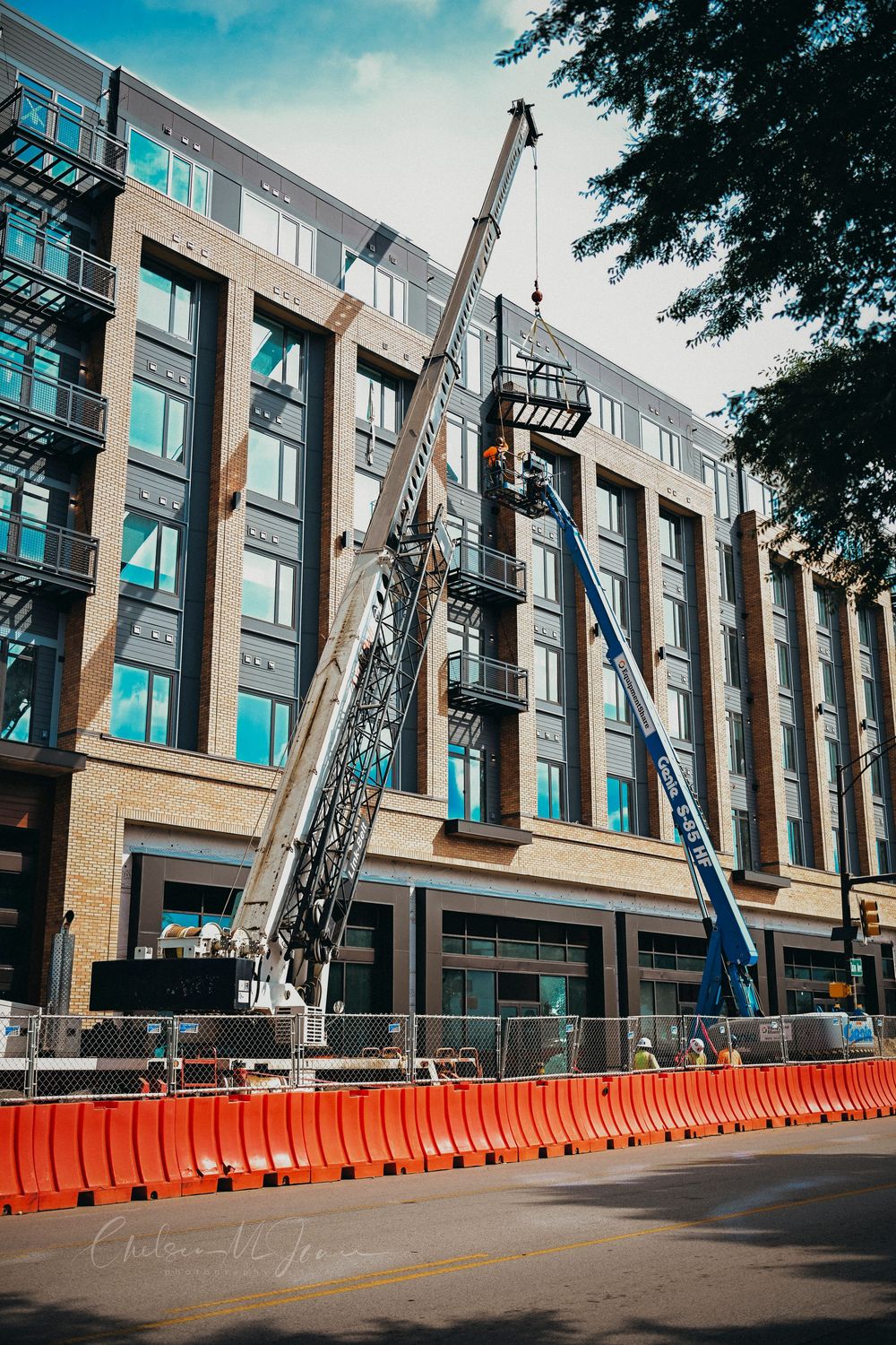 Construction cranes and orange barriers surround a modern multi-story brick building during urban renovation work on a city street.
