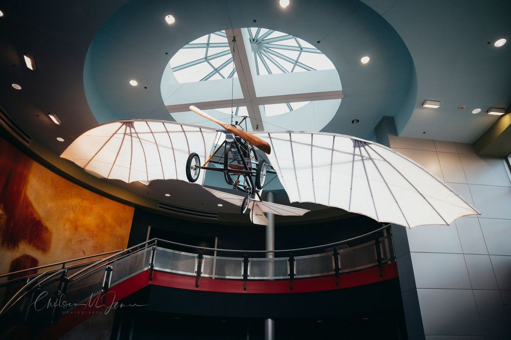 Vintage glider aircraft display hanging in a modern museum atrium with circular skylight and curved balcony.