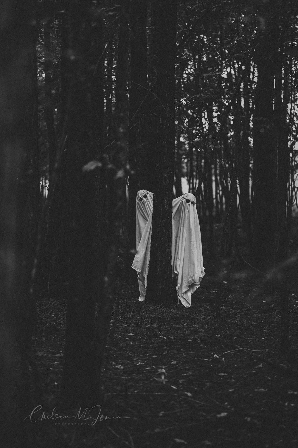 Two ghostly white-draped figures float eerily among dark forest trees in a haunting black and white fine art photograph.