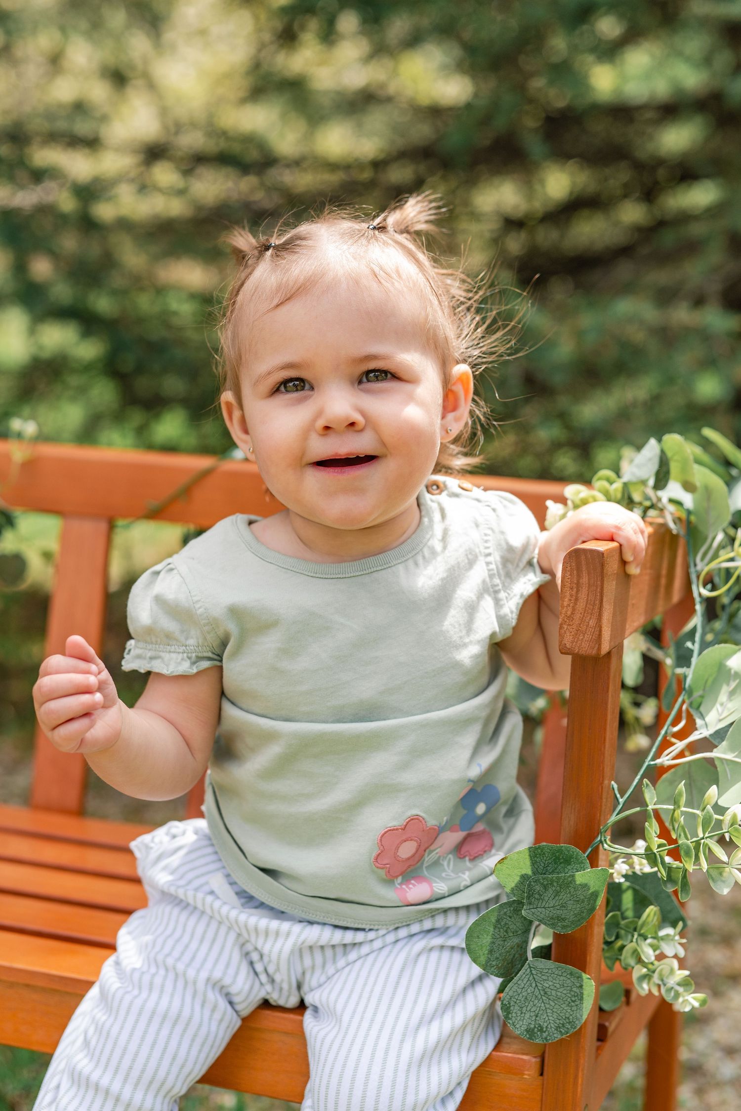 A toddler girl in a purple gingham dress stands outdoors near dark red bushes, smiling with her hand near her mouth.