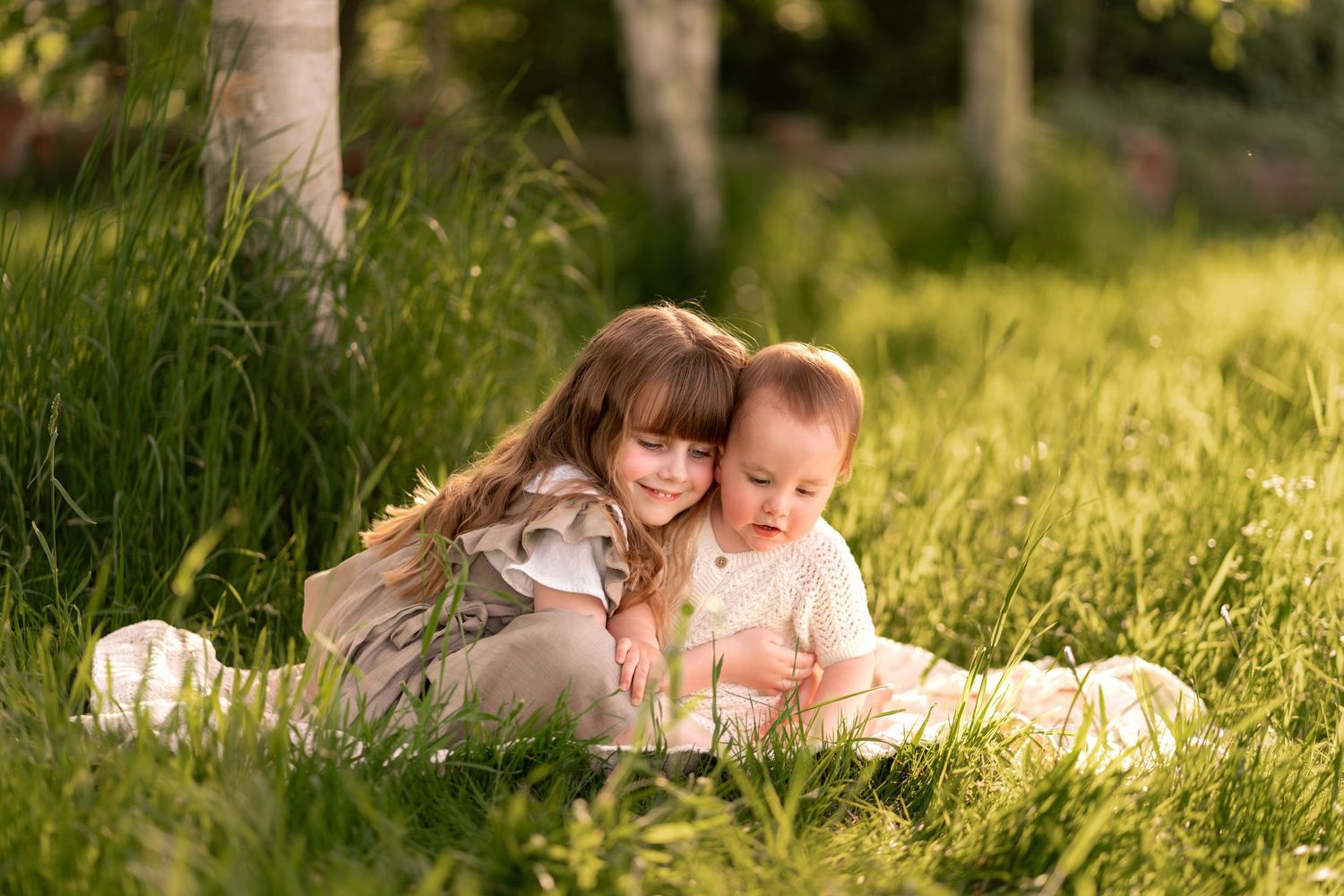 Two young children sitting in lush green grass outdoors, older girl hugging baby sibling in warm golden sunlight.