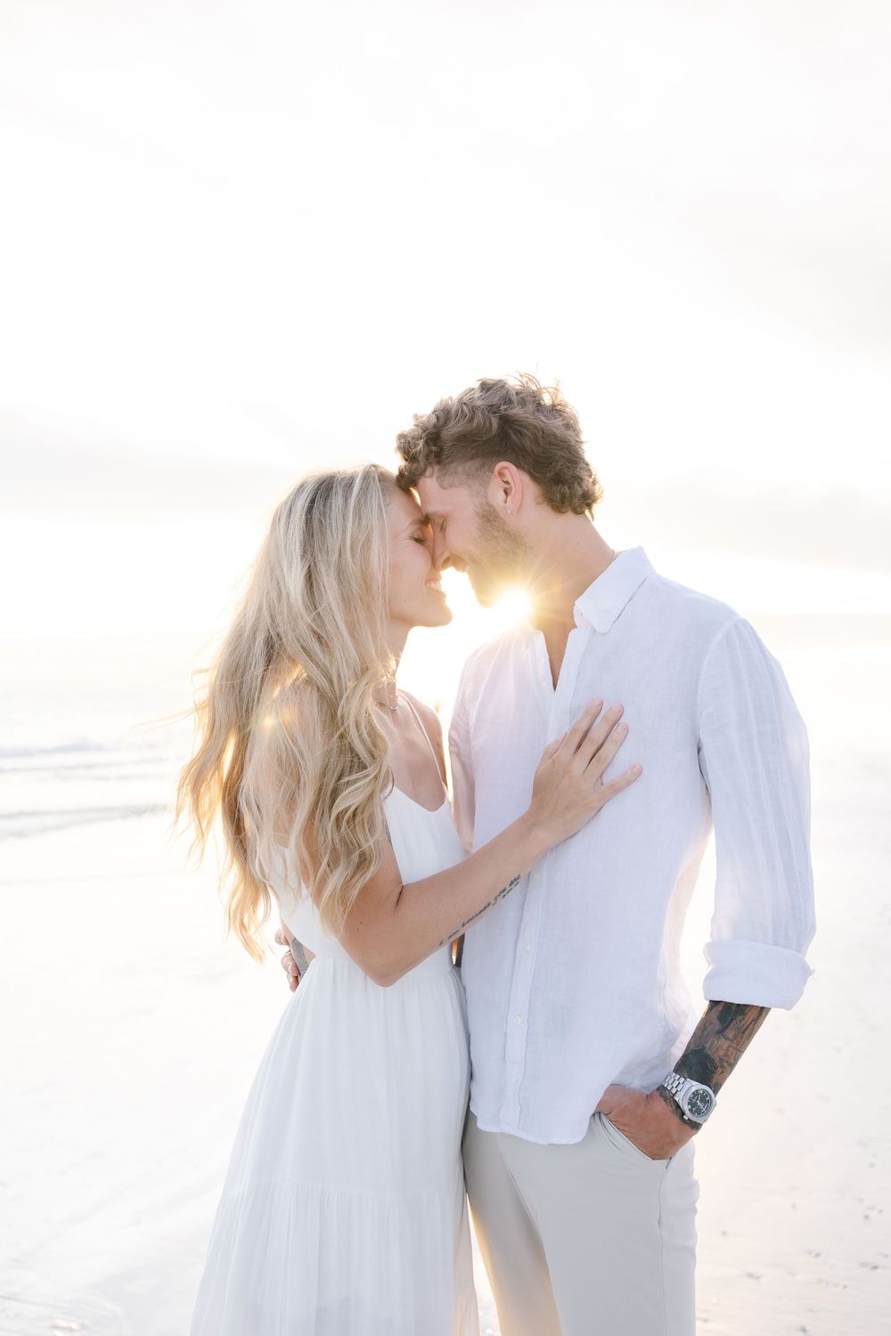 Couple sharing a romantic moment on a sunlit beach, dressed in white, with golden backlight and ocean waves behind them.