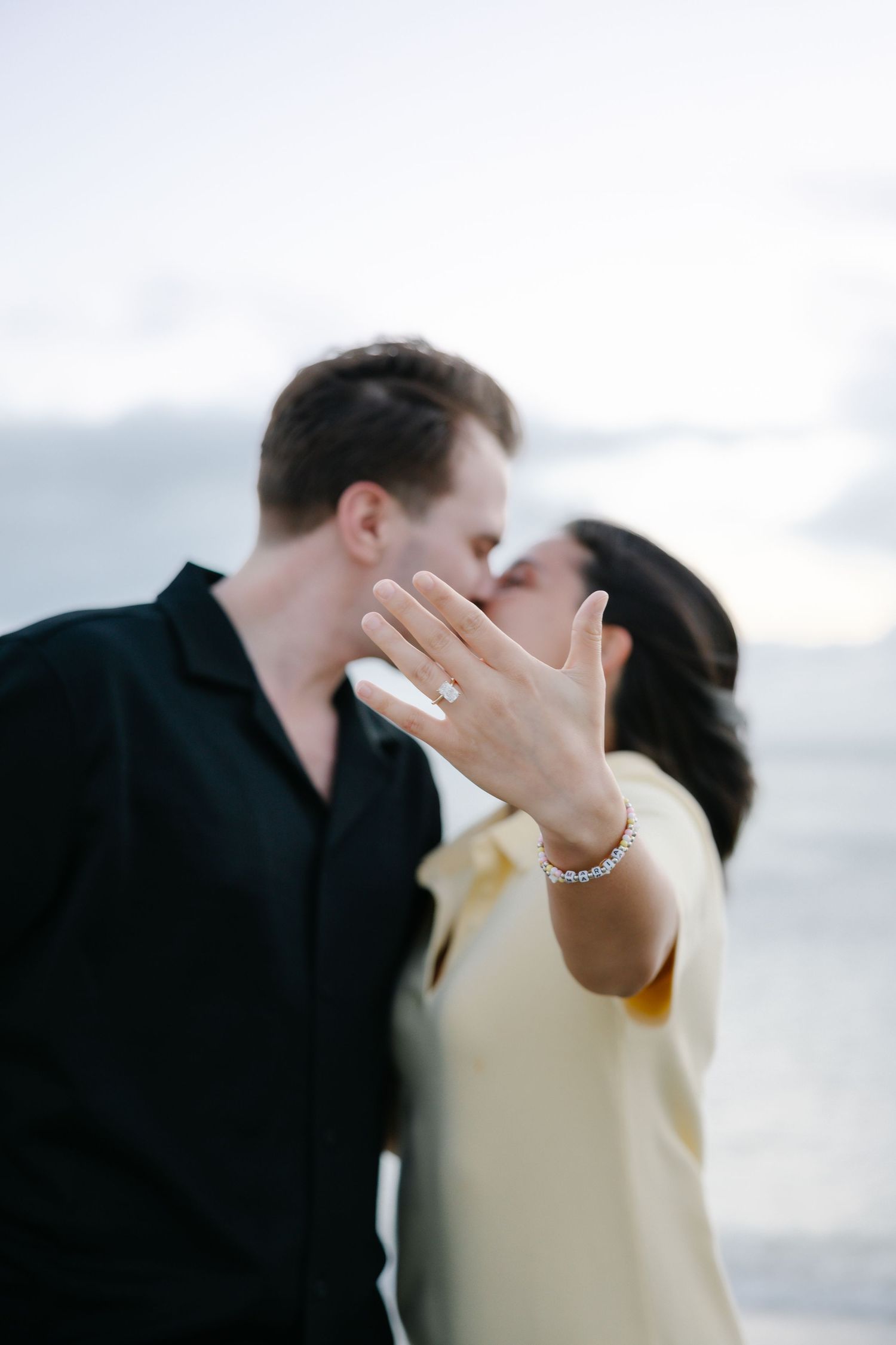 Newly engaged couple kissing on beach while woman shows off diamond ring.