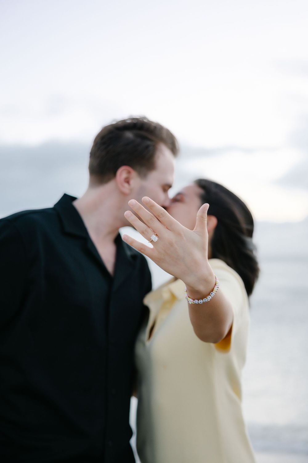 Newly engaged couple kissing on beach while woman shows off diamond ring.