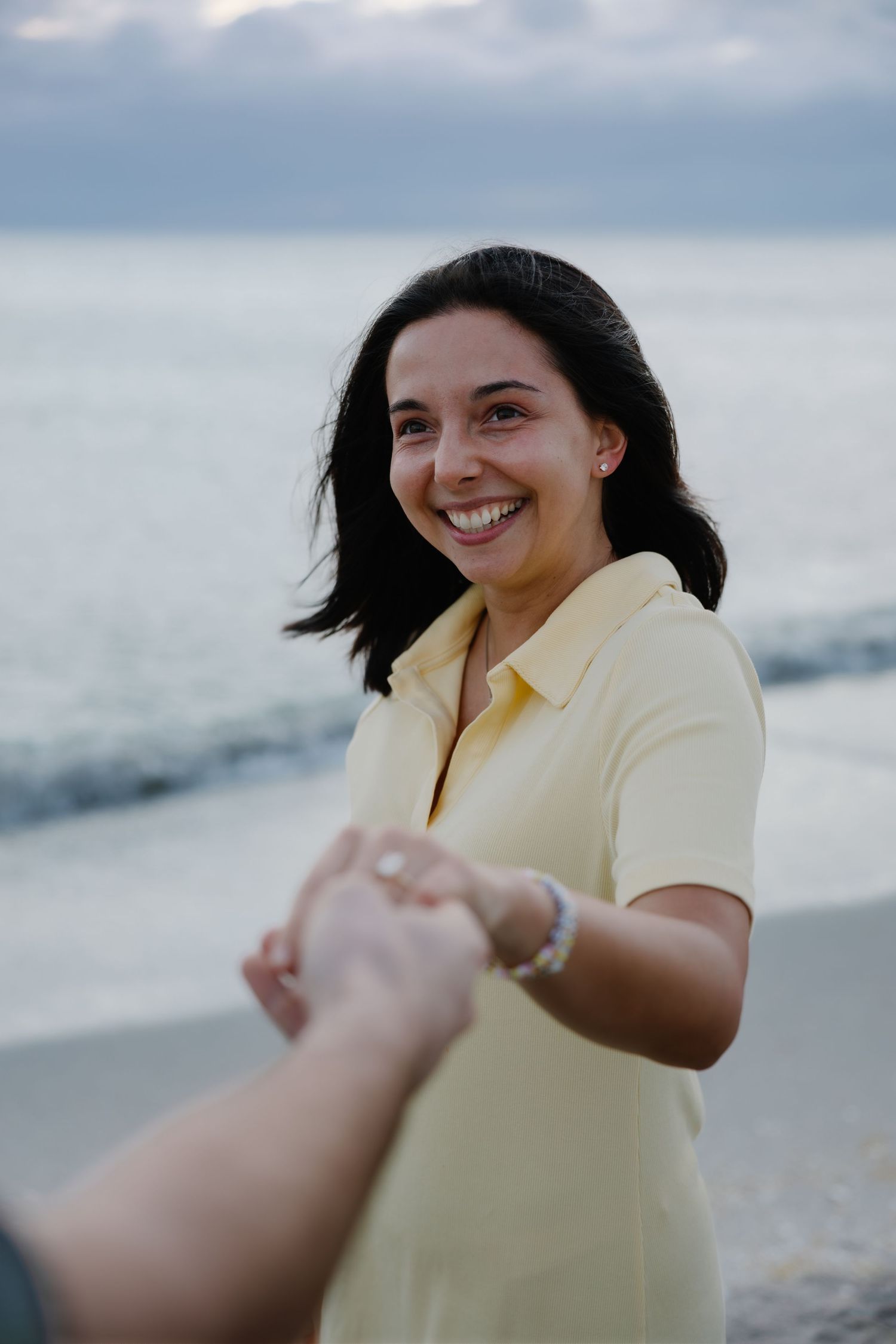 Smiling dark-haired woman in yellow polo holding hands on beach at dusk.