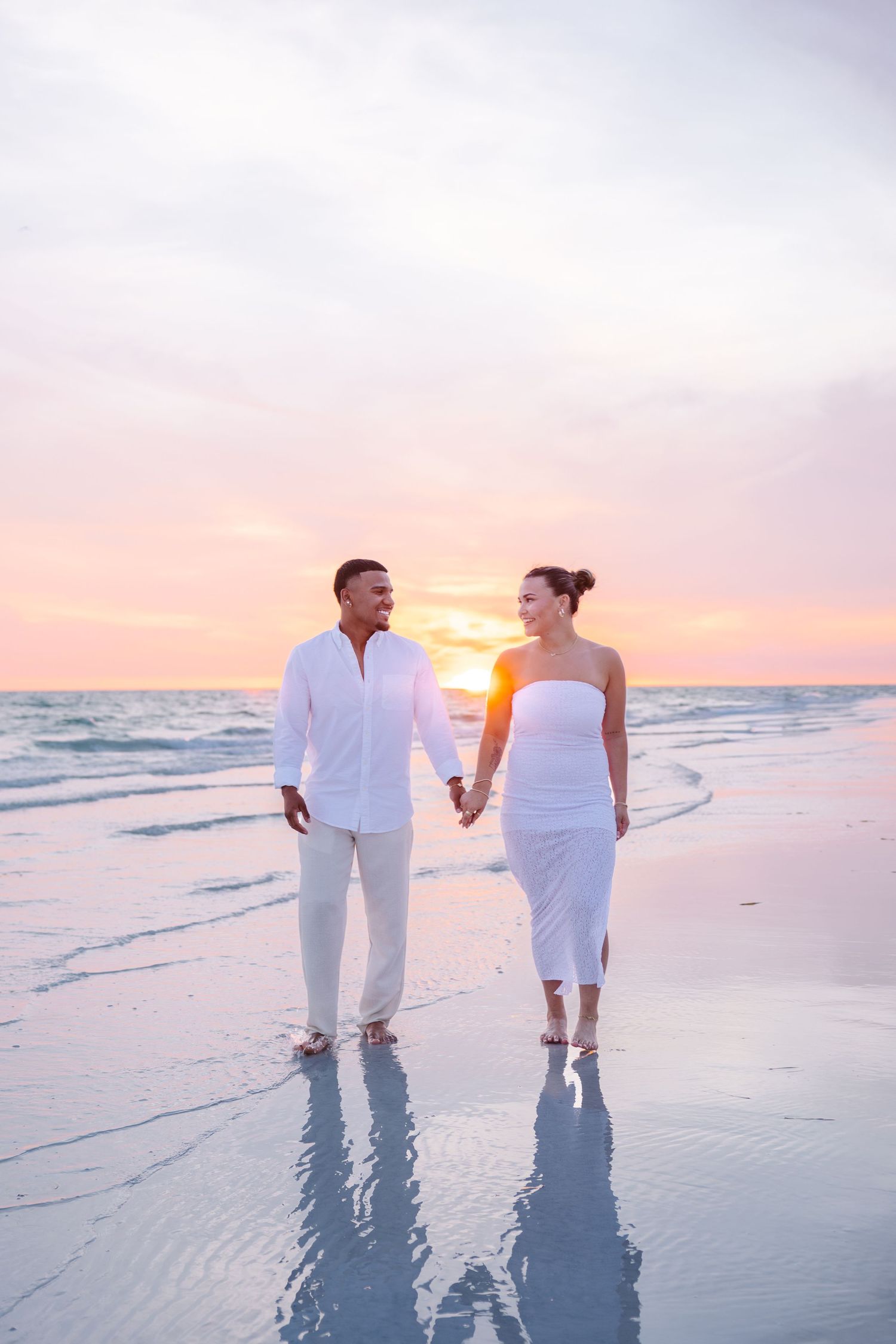 A couple in matching white outfits walks hand in hand on a beach at vibrant sunset.