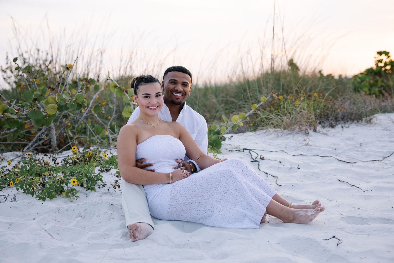 A smiling interracial couple sits together on white sand beach at dusk in white outfits.
