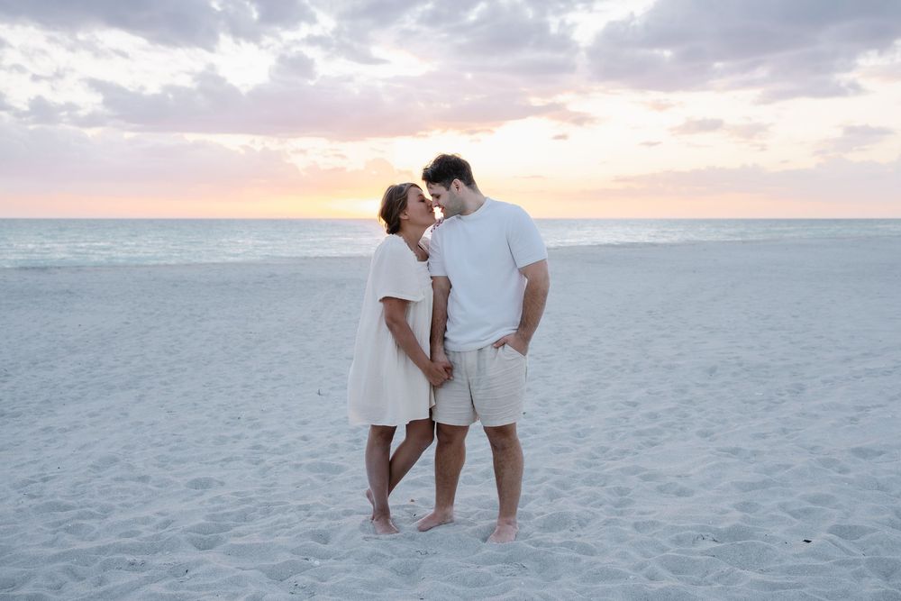 Romantic couple in white outfits sharing a kiss on sandy beach at sunset.
