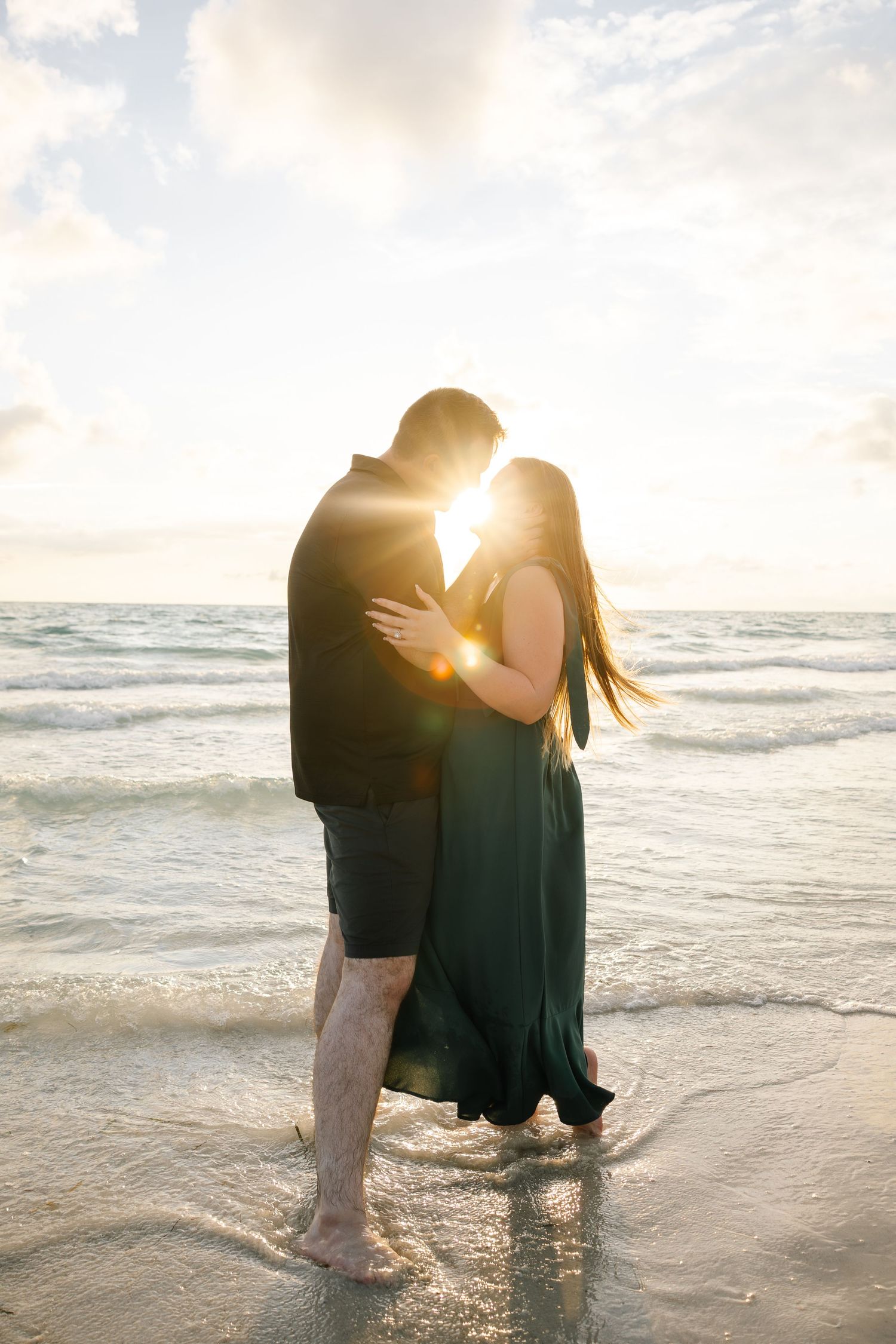 Couple embracing on beach at golden sunset with sun flare between them.