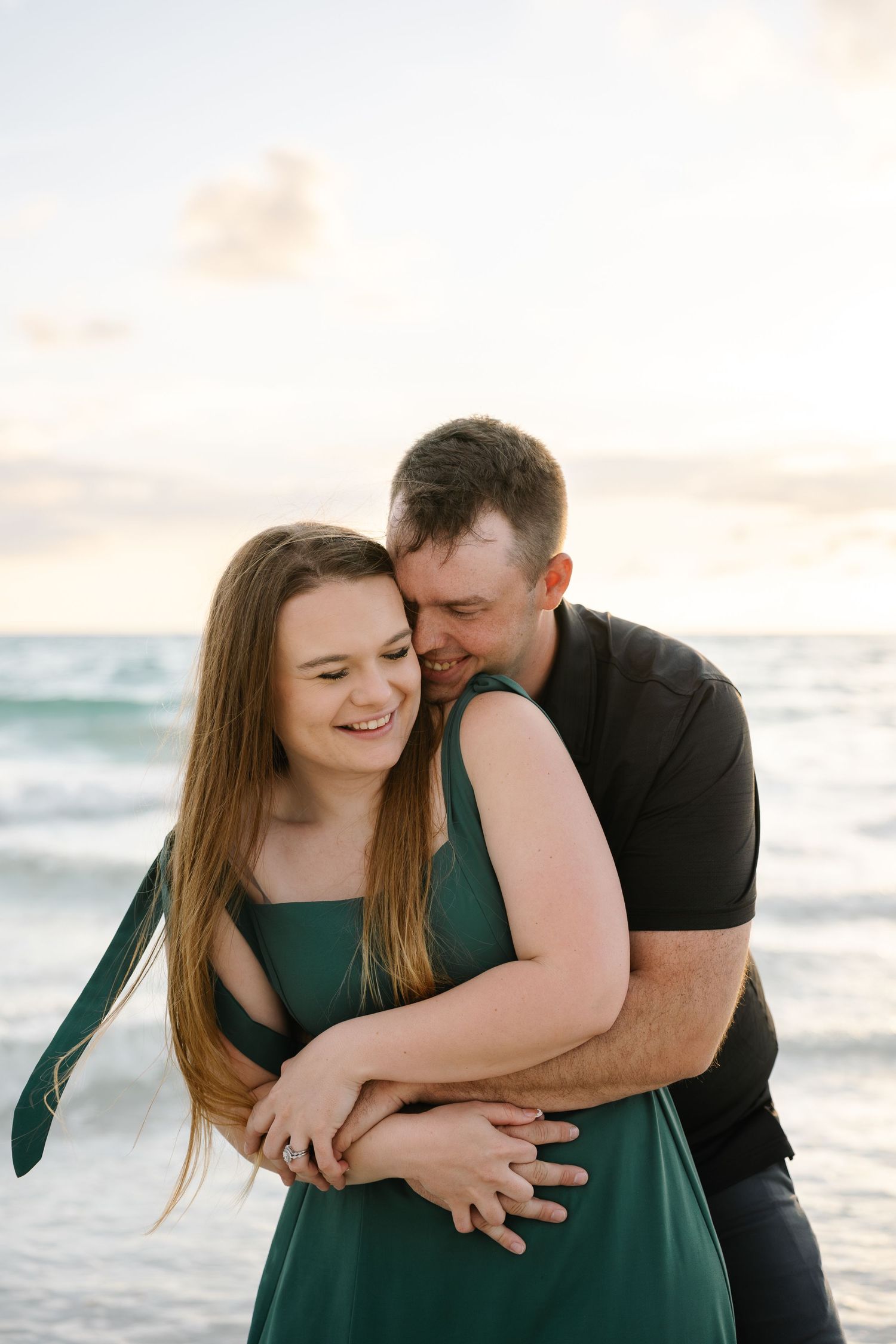 Man in black shirt hugging smiling woman in green dress on beach at sunset.