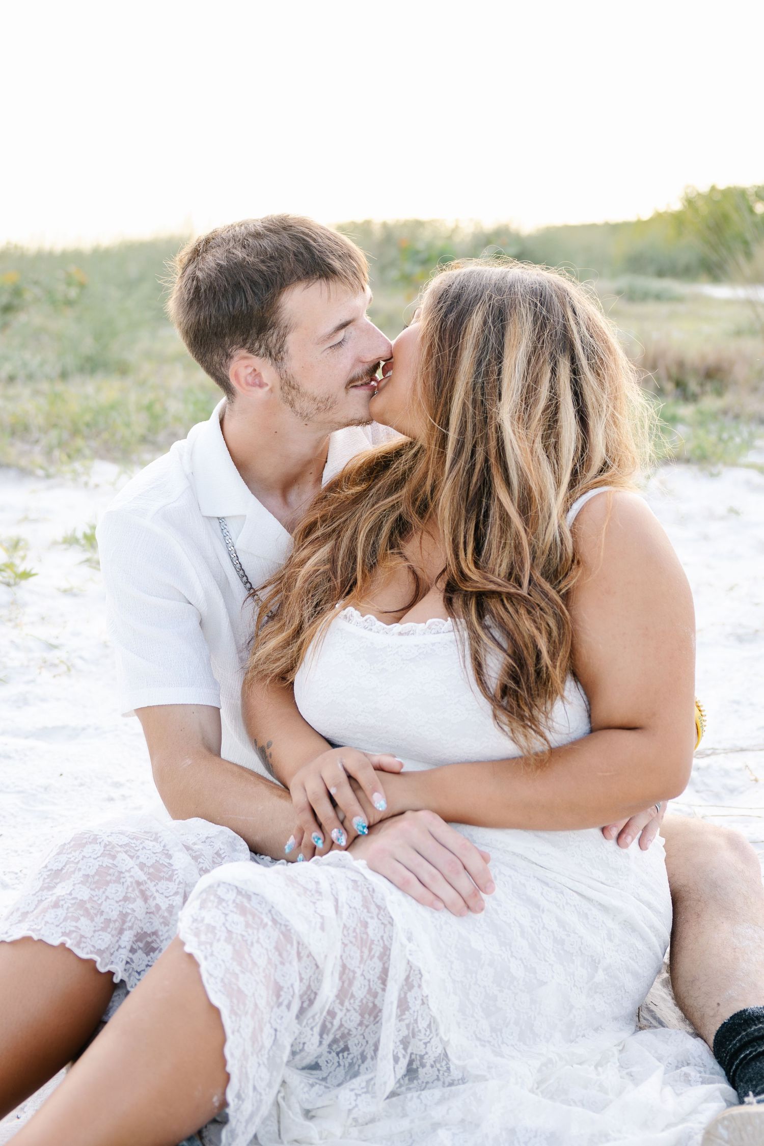 A young couple shares a kiss while sitting on sandy beach dunes at golden hour.