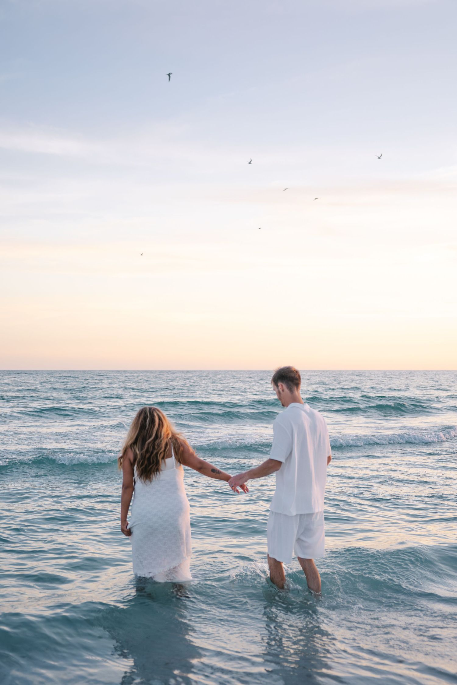 Couple in white outfits holding hands while wading in ocean waves at sunset.