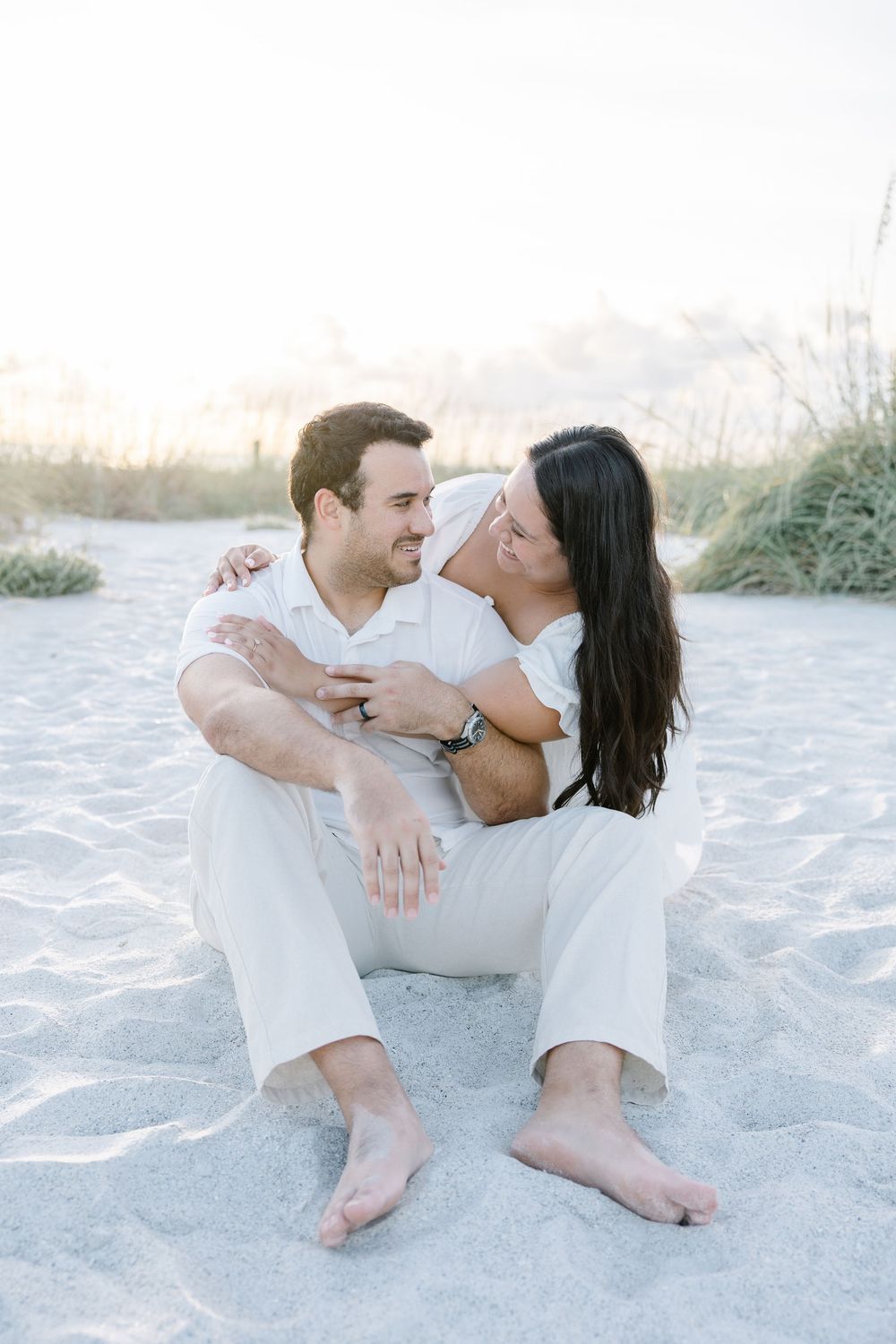 A couple in white outfits sits barefoot on sandy beach, sharing a romantic moment at sunset.