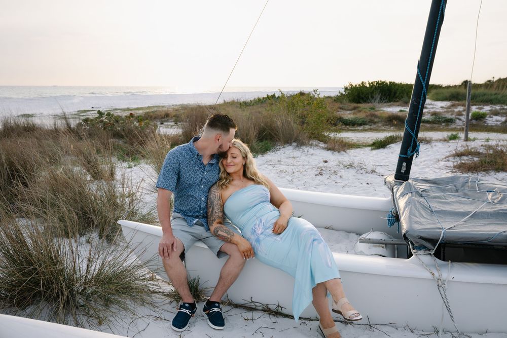 Couple sitting together on beached sailboat surrounded by dunes at sunset.