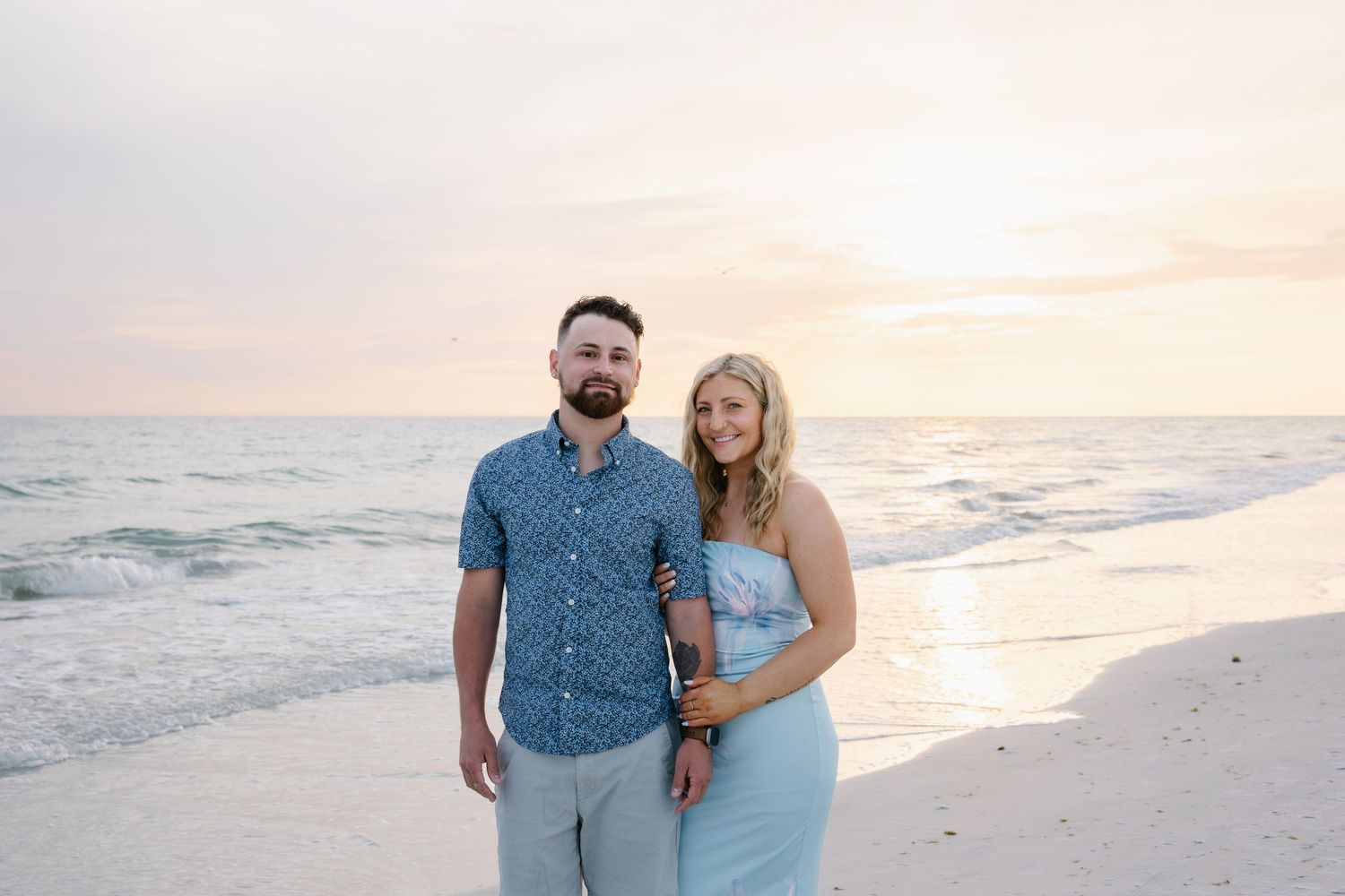Smiling couple in coordinating blue outfits posing on beach at pink sunset.