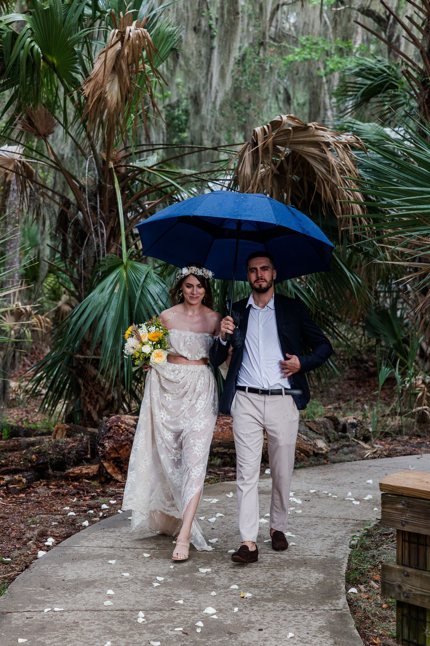 Bride and groom walking under blue umbrella on rainy wedding day surrounded by lush tropical greenery.