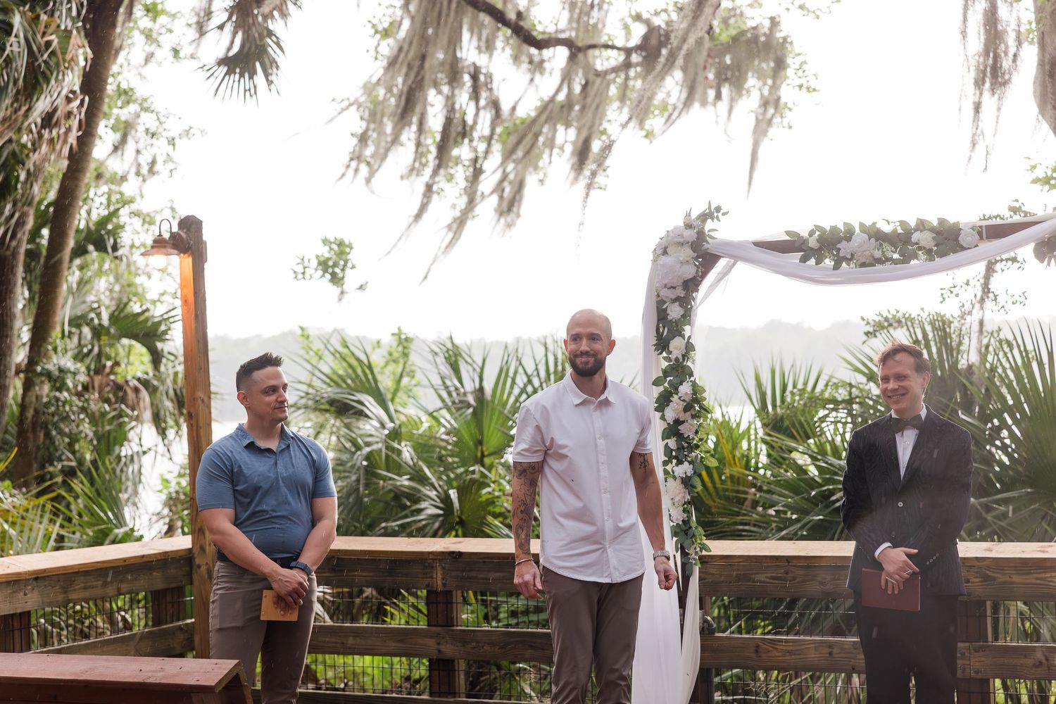 Three men stand on a wooden deck at an outdoor wedding ceremony surrounded by lush tropical greenery.