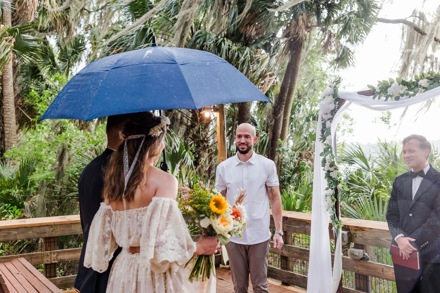Groom smiles as bride approaches under blue umbrella at outdoor tropical wedding ceremony on wooden deck.
