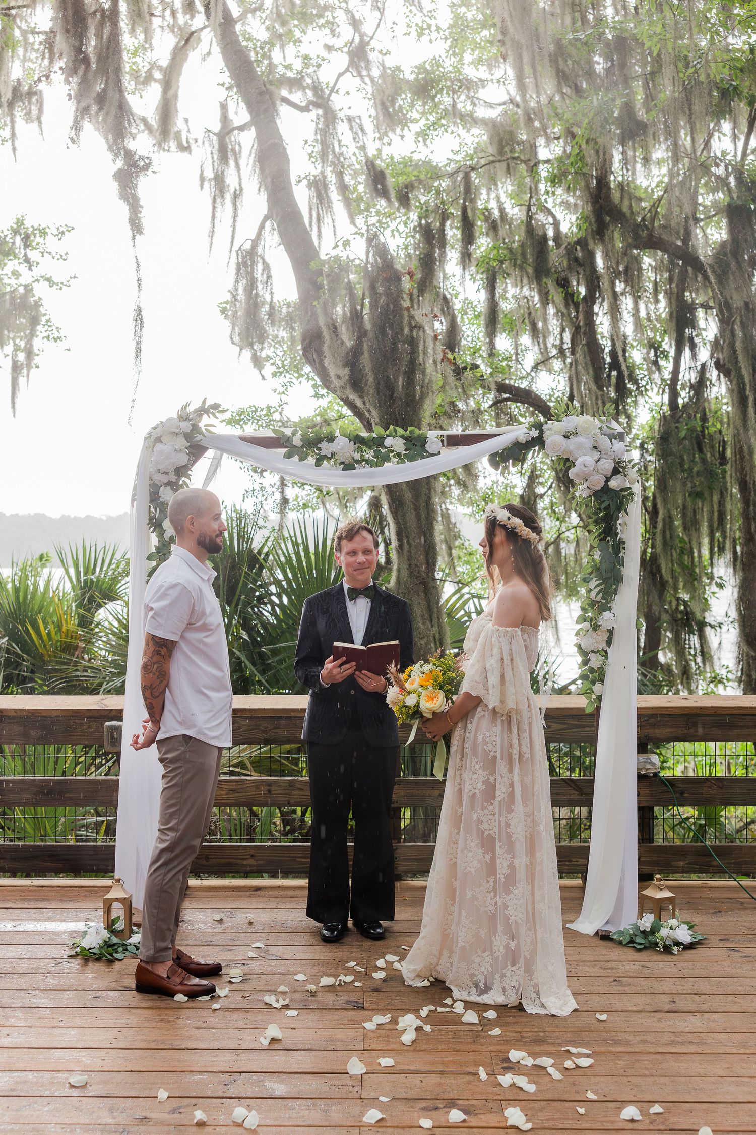 Couple exchanges vows under floral arch on wooden deck with Spanish moss trees and waterfront backdrop.