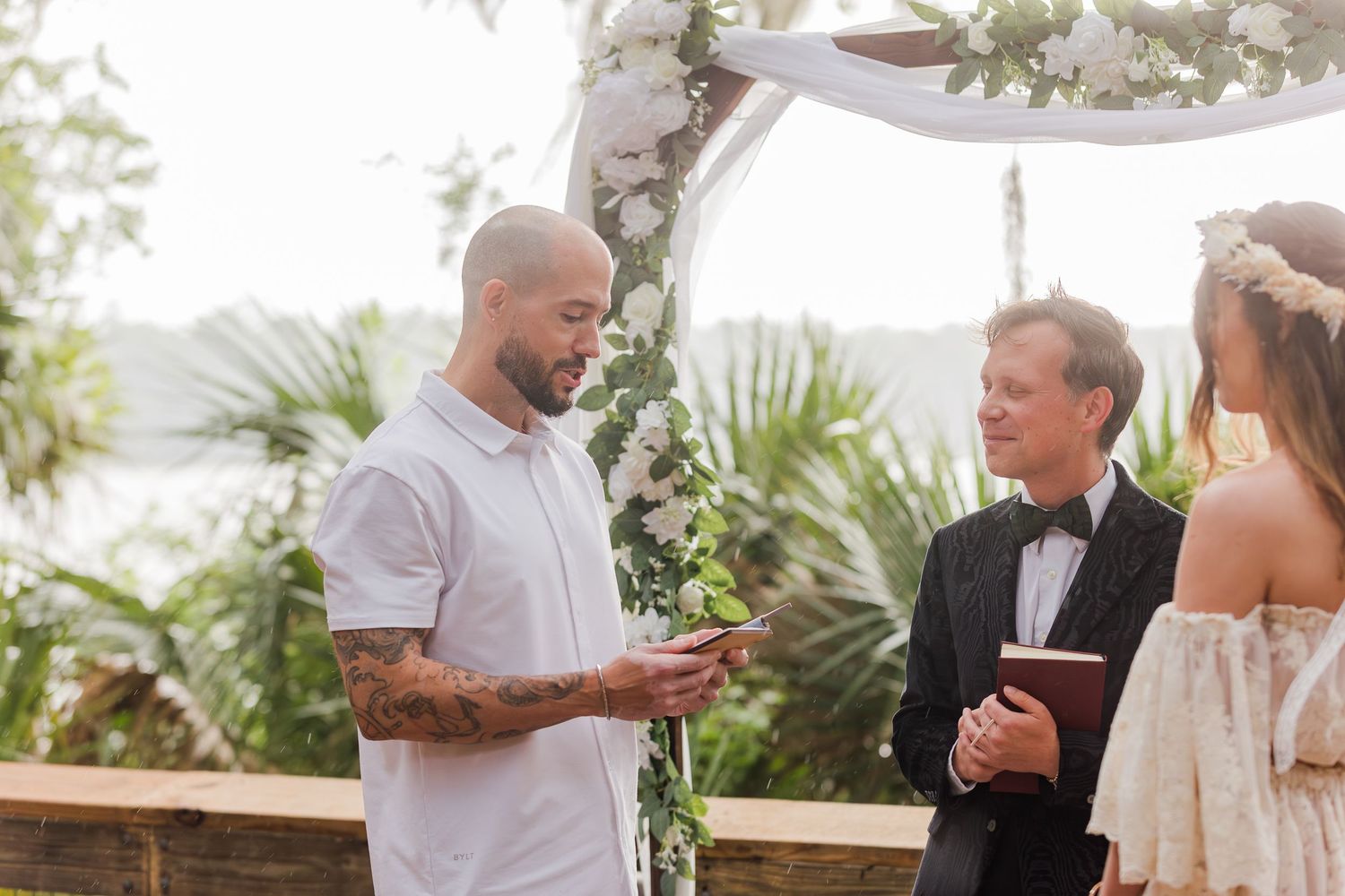Groom reads vows at outdoor wedding ceremony with floral arch, officiant, and bride in white dress near waterfront.