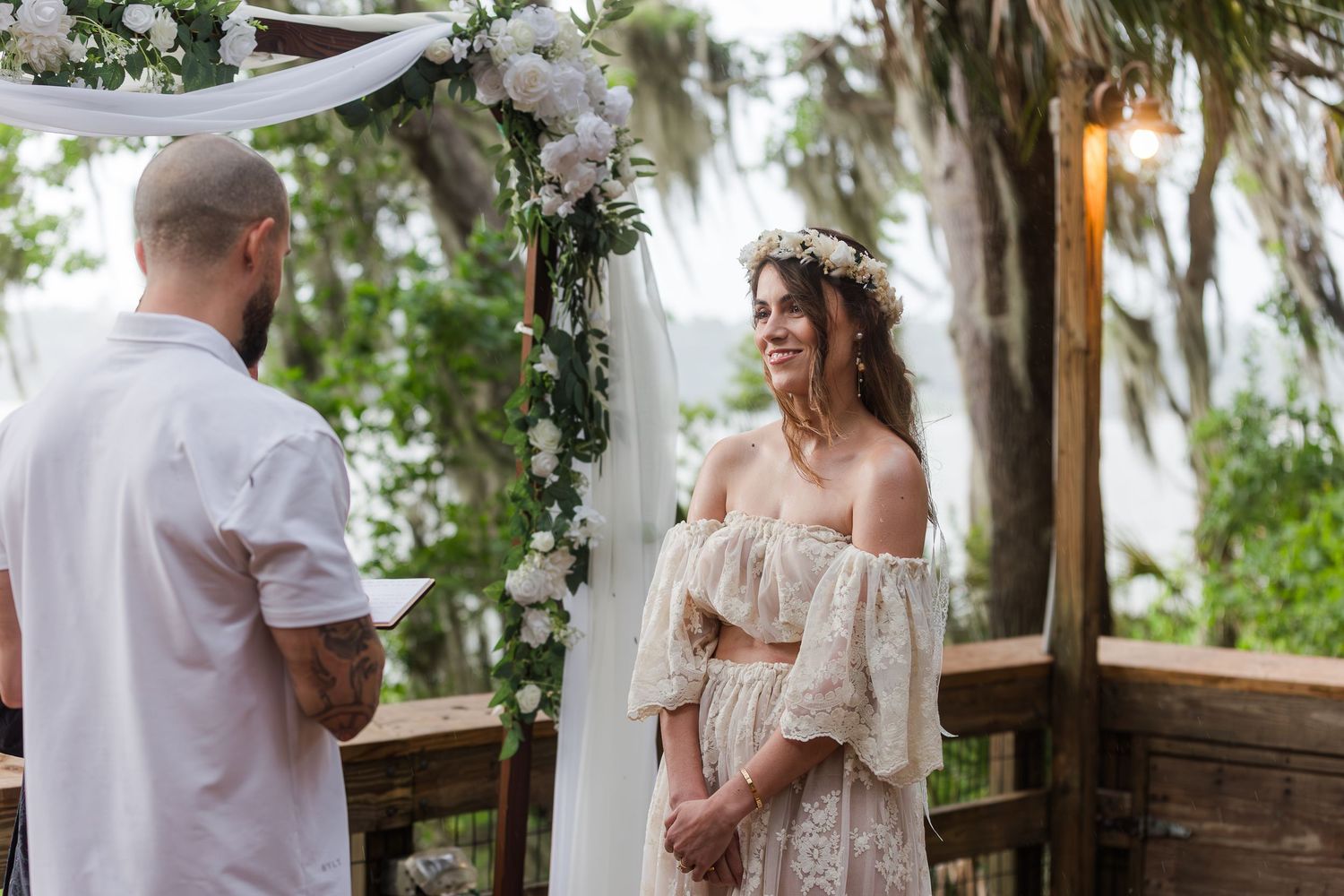 Bride in lace off-shoulder gown smiles at groom during outdoor wedding ceremony under floral arch.