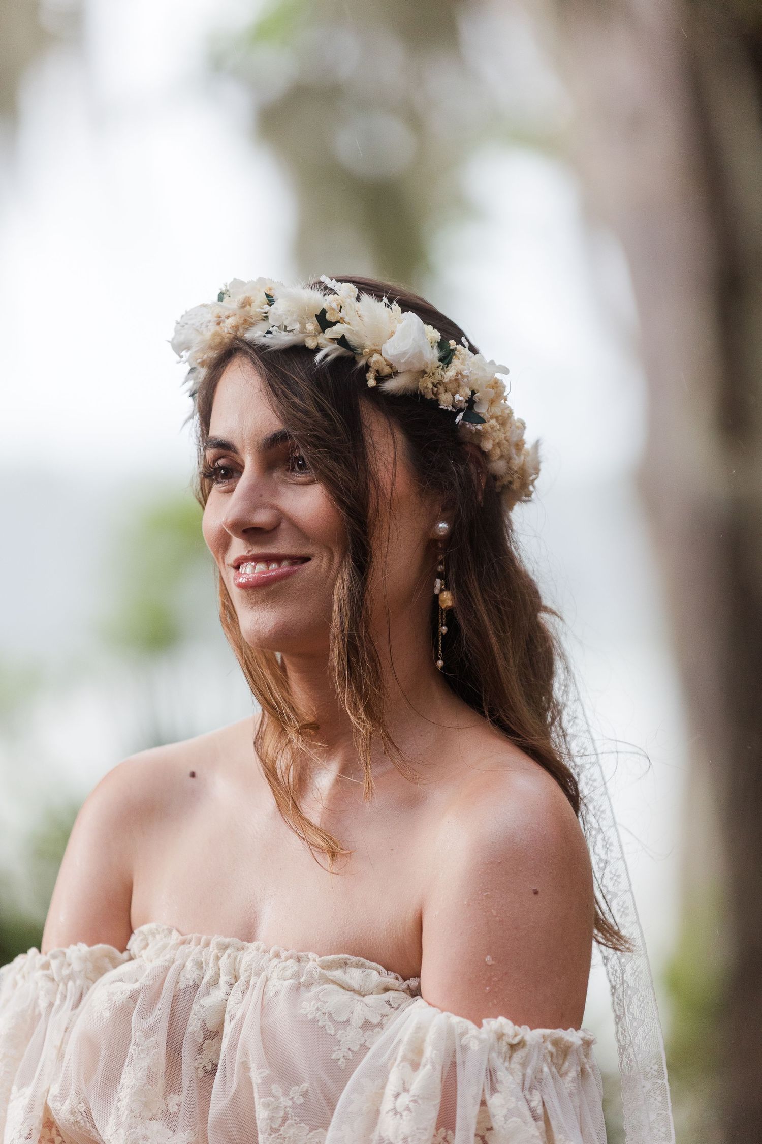 Smiling bride wearing a white floral crown and off-shoulder lace wedding dress with pearl earrings outdoors.