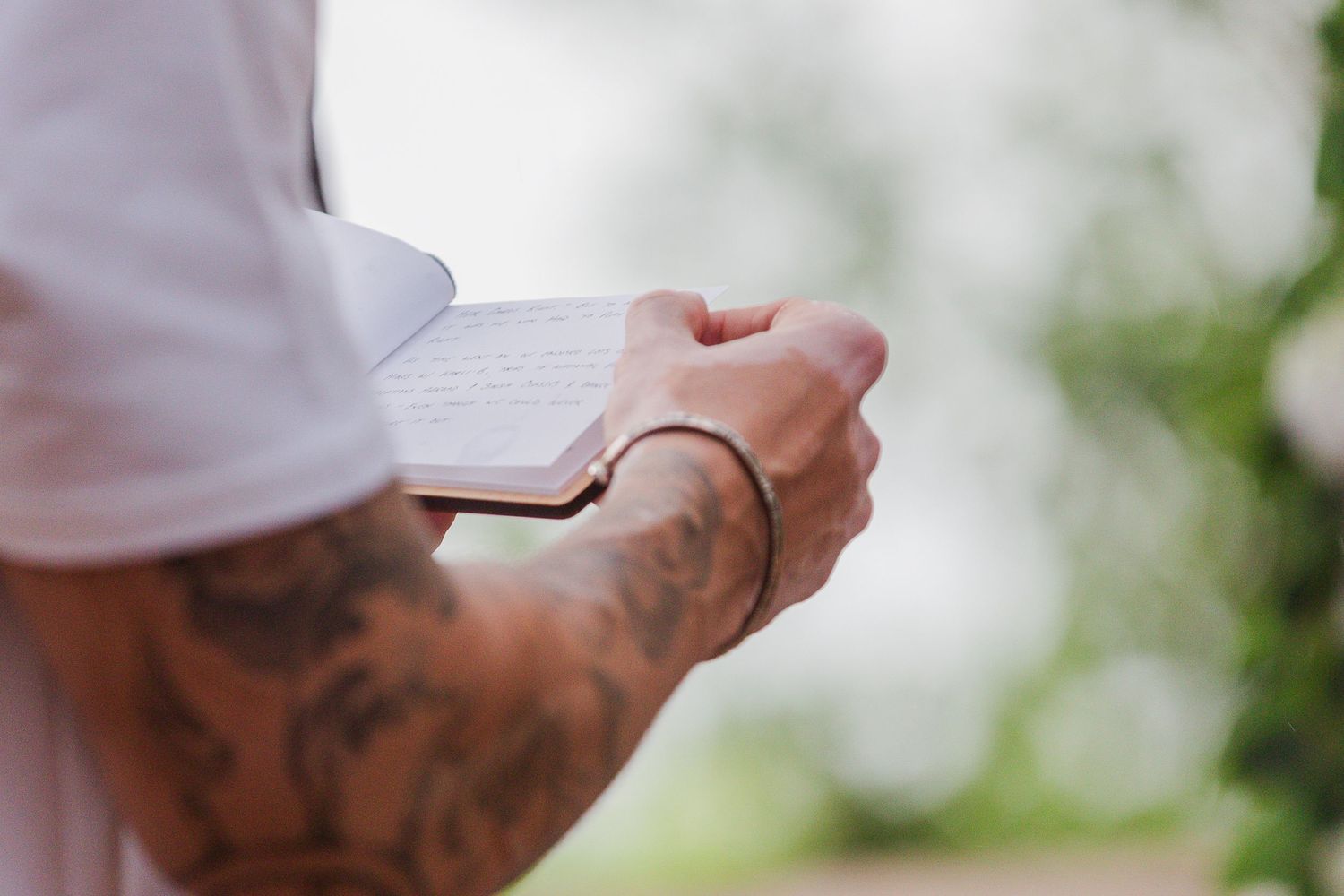 Tattooed man holding a small open notebook outdoors, wearing a white shirt and black bracelet.