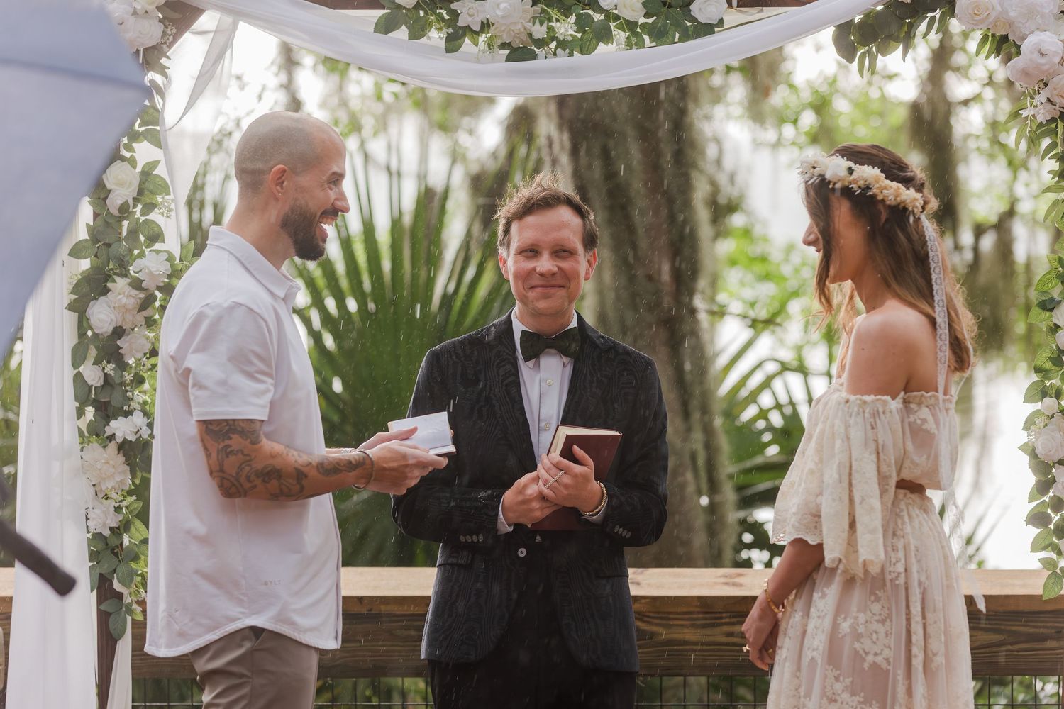 Outdoor wedding ceremony with tattooed groom, officiant in tuxedo, and bride in floral crown under white flower arch.
