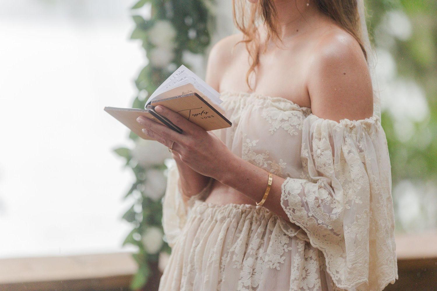 Woman in floral off-shoulder dress reading a notebook outdoors, wearing gold bracelets near lush greenery.