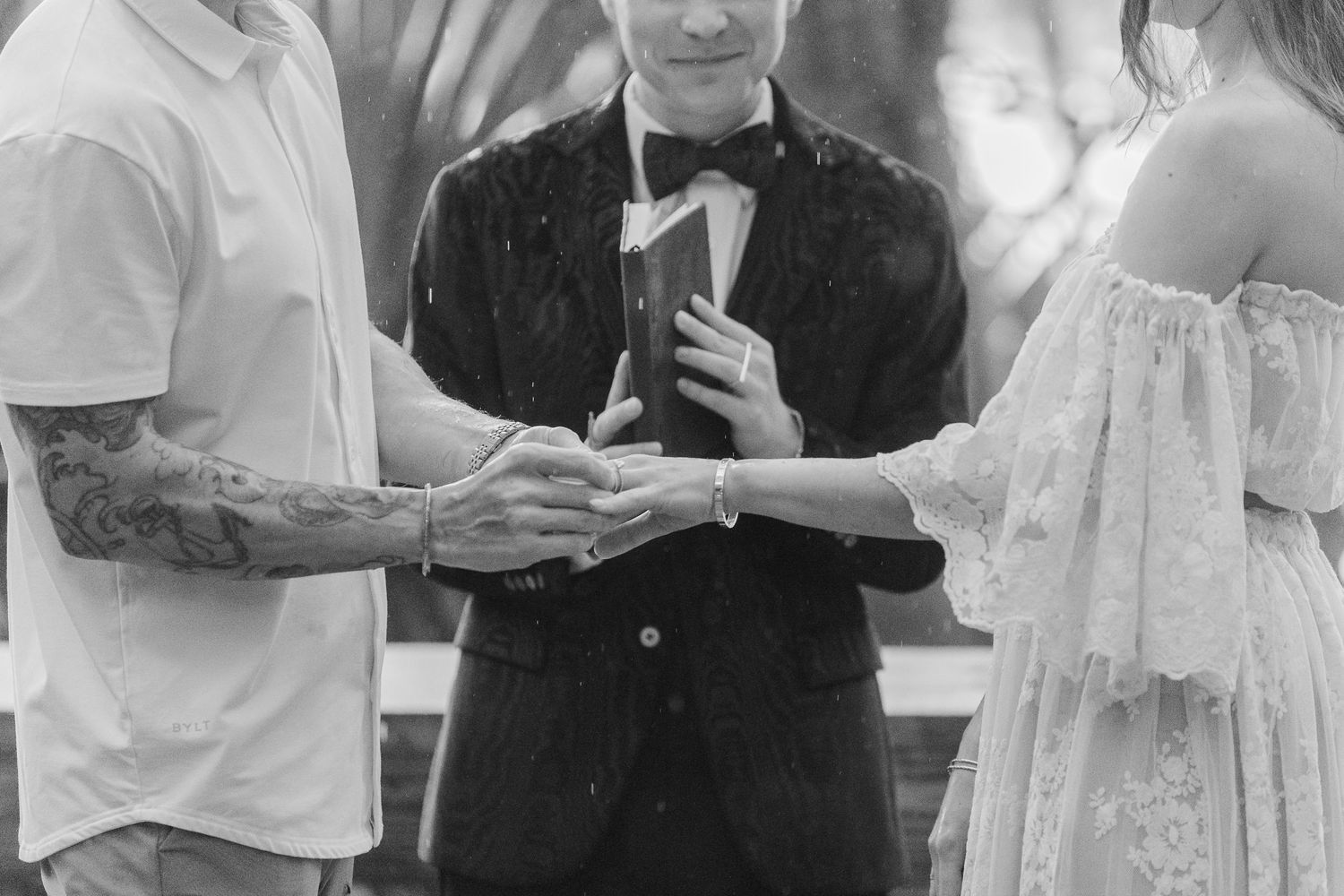 Black and white wedding ceremony photo of couple exchanging vows with officiant holding Bible outdoors.