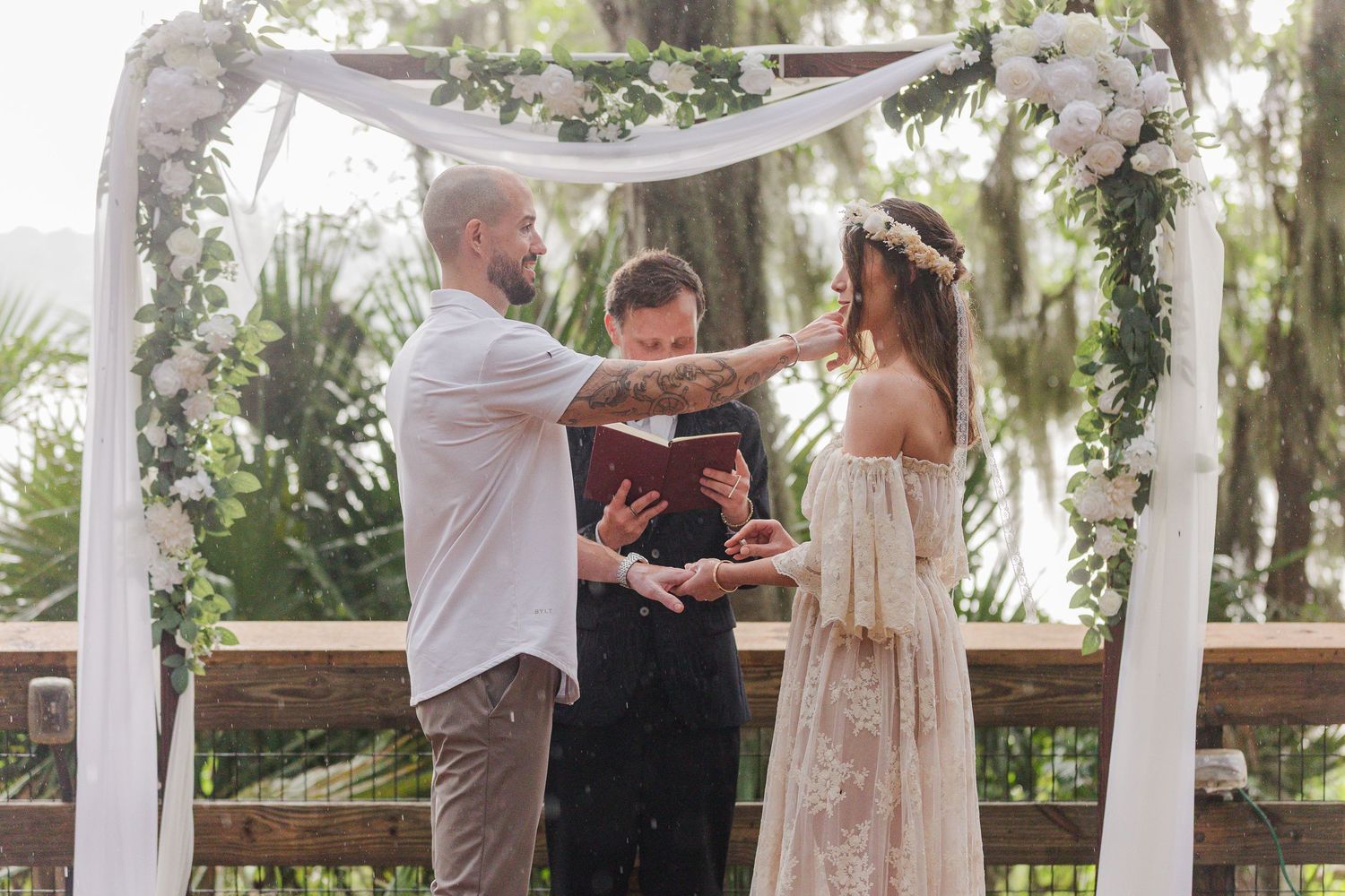 Outdoor wedding ceremony under floral arch with bride, groom, and officiant on wooden deck surrounded by lush greenery.