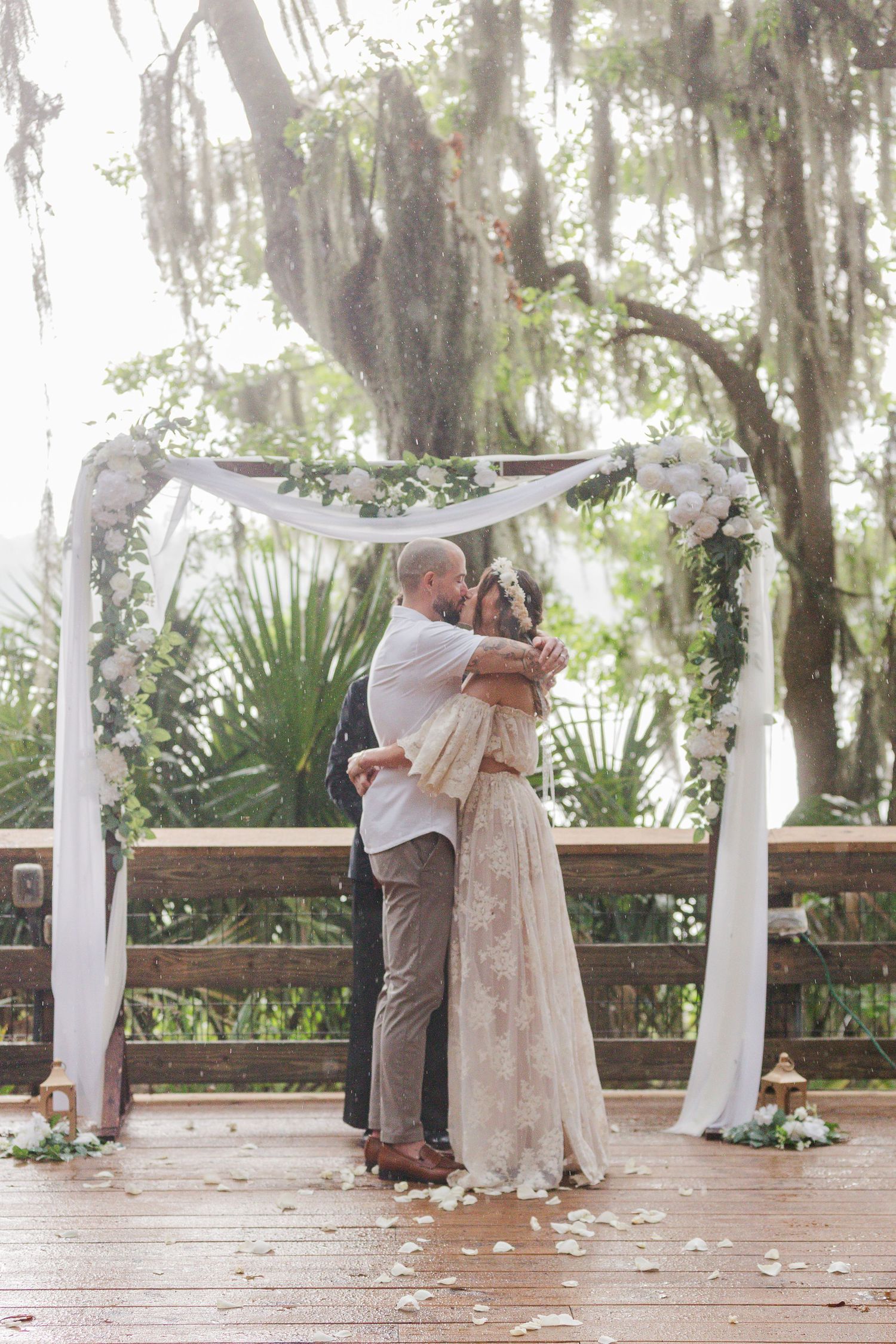 Couple embracing under a floral wedding arch on a wooden deck surrounded by lush greenery and white rose petals.