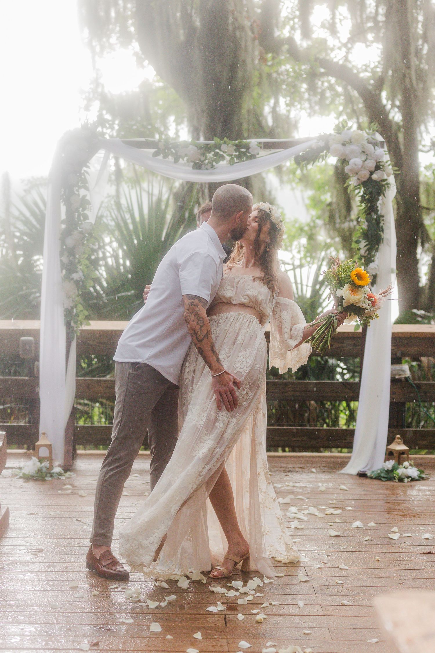 Couple sharing a kiss under a floral arch on a wooden deck during an outdoor wedding ceremony.