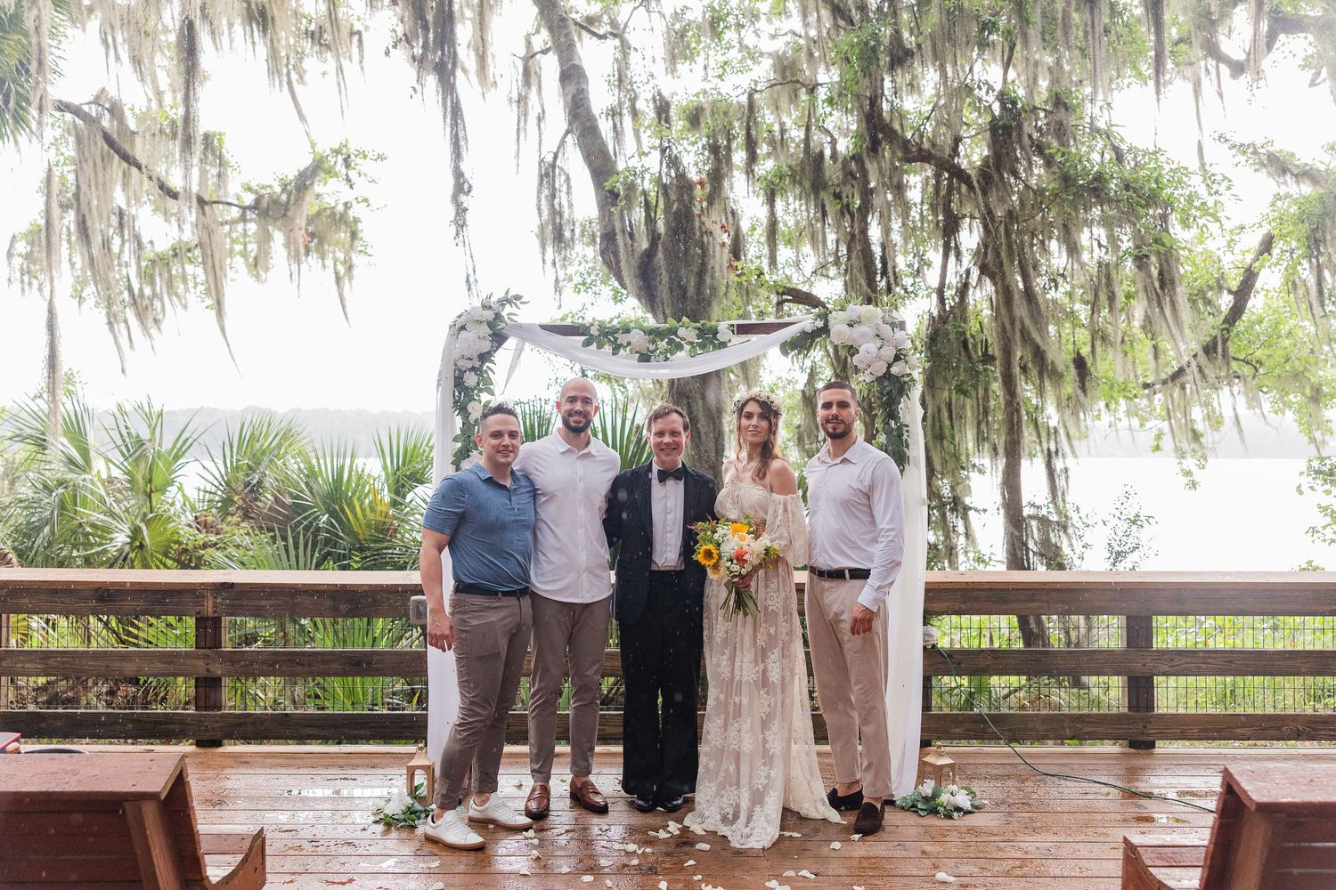 Five guests pose under a floral arch on a wooden deck at an outdoor wedding ceremony with Spanish moss trees.
