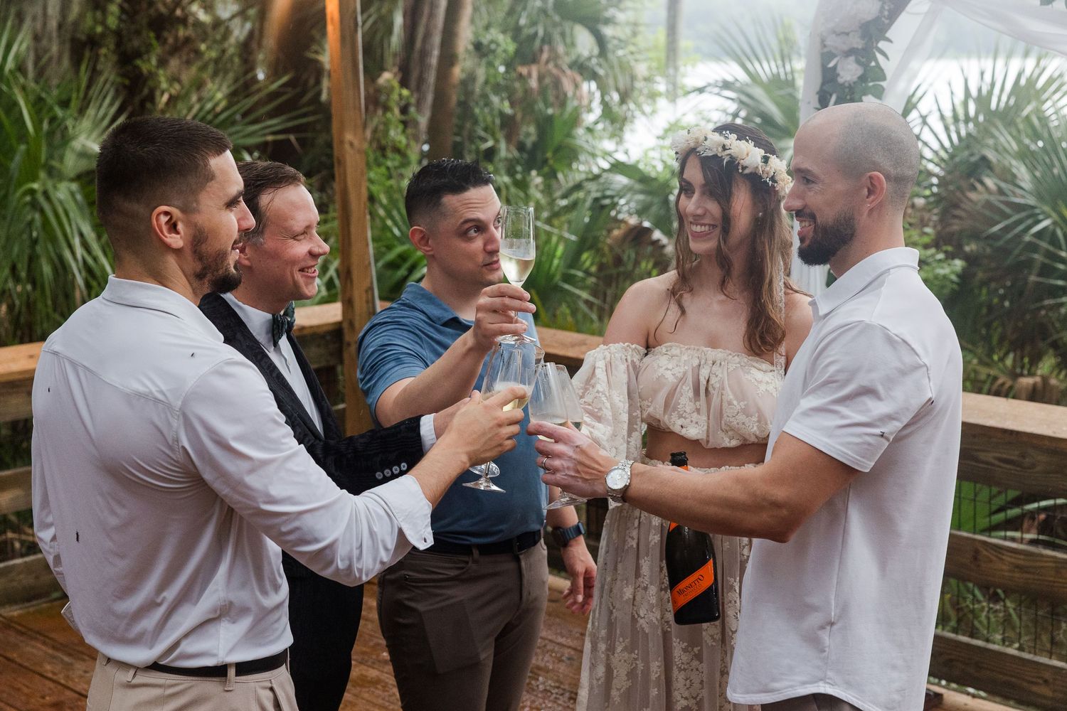 Group of five people toasting with champagne at an outdoor wedding celebration surrounded by tropical greenery.