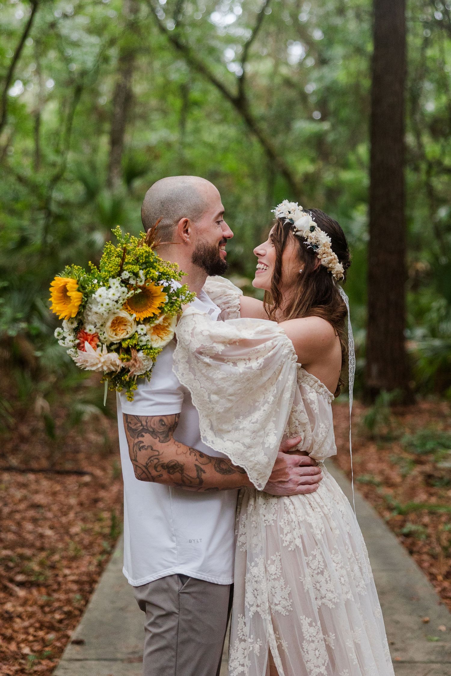 Bride in lace dress with floral crown embraces tattooed groom holding sunflower bouquet on forest path.