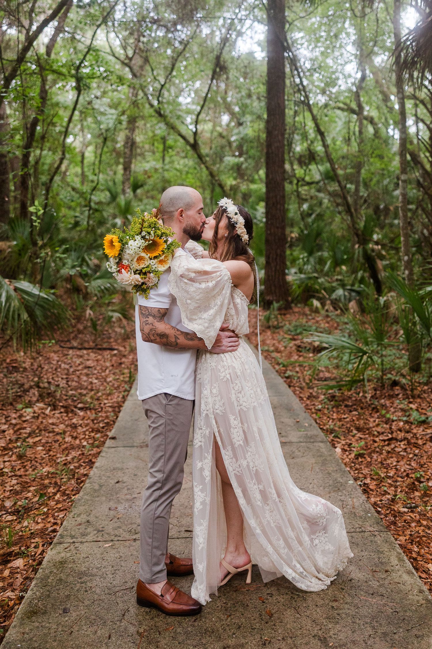 Bride in lace boho gown with floral crown kisses tattooed groom holding sunflower bouquet on forest path.
