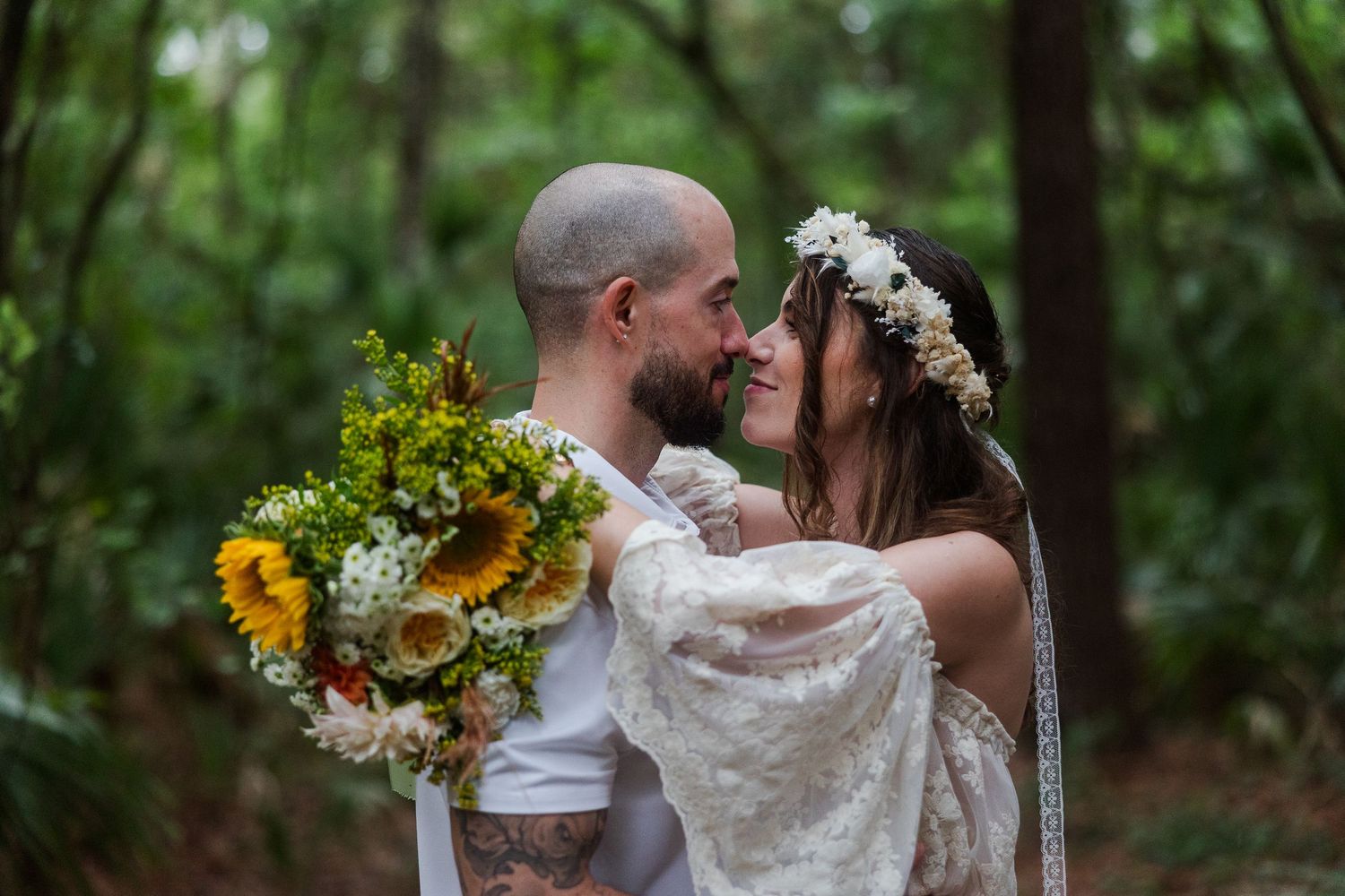 Bride and groom share a romantic moment in lush forest setting, bride holding sunflower bouquet wearing floral crown.