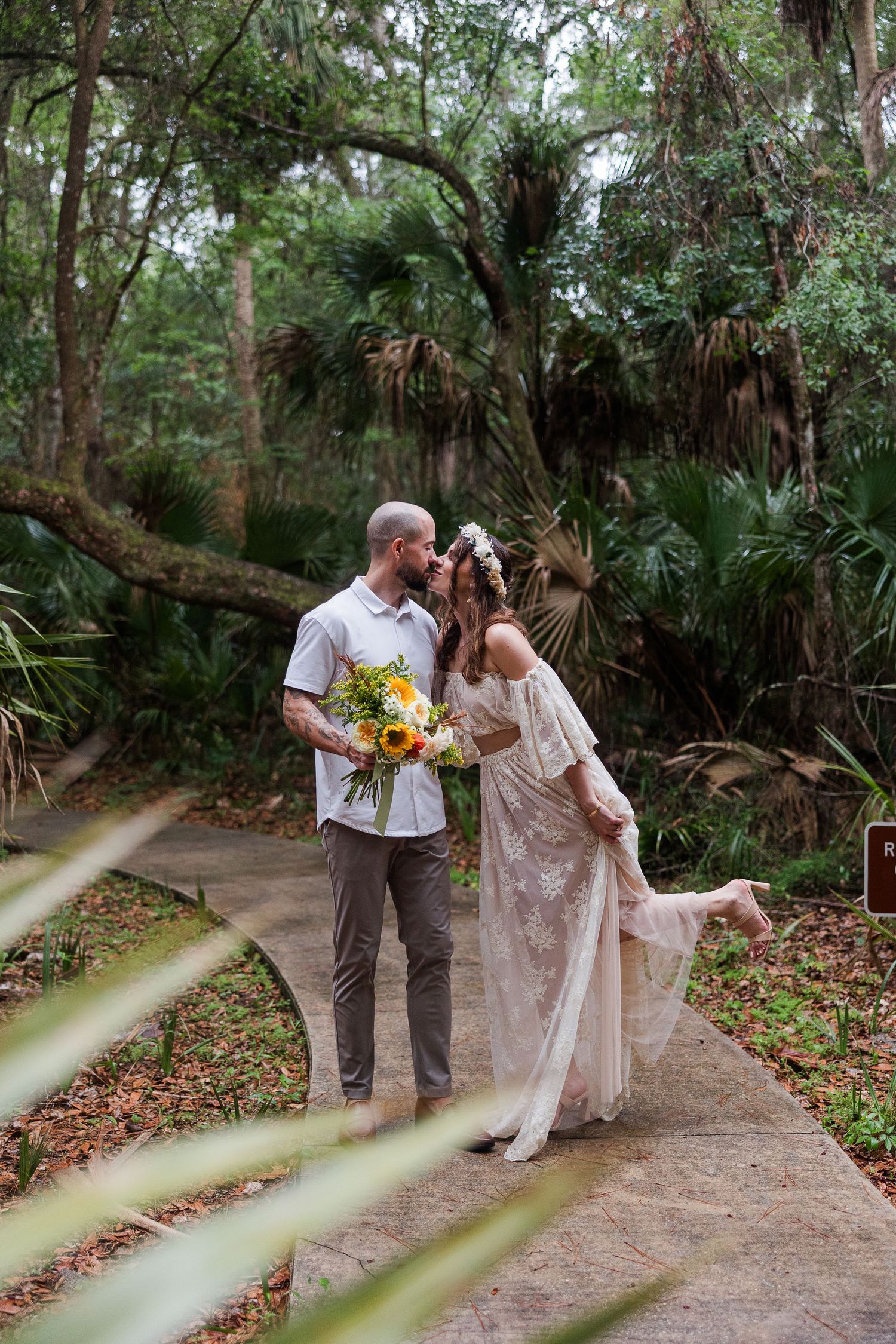 Couple shares romantic kiss on woodland path, bride in off-shoulder lace dress holding sunflower bouquet.