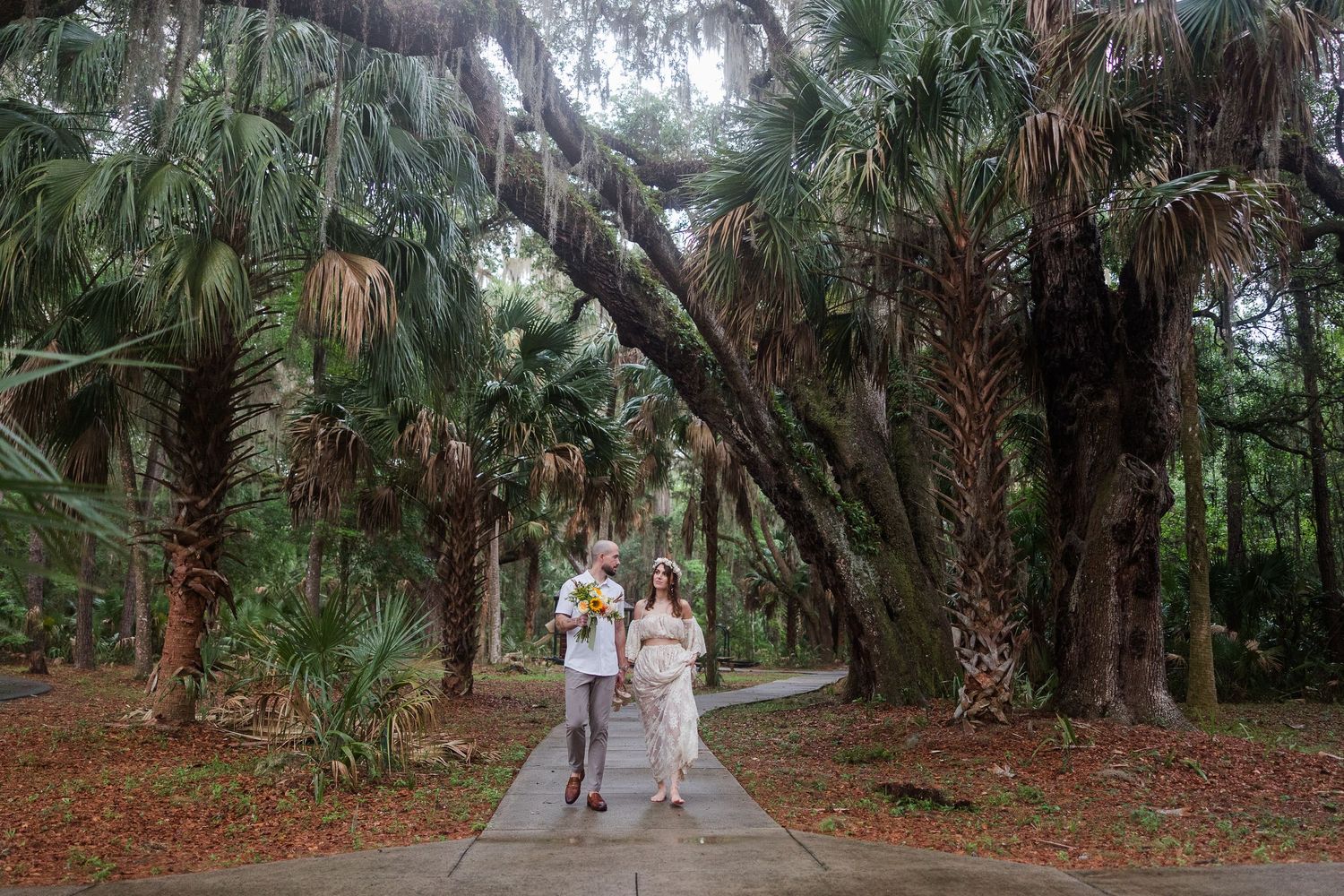 Couple walks hand-in-hand under sprawling oak and palm trees on a misty path, woman holding yellow flowers.