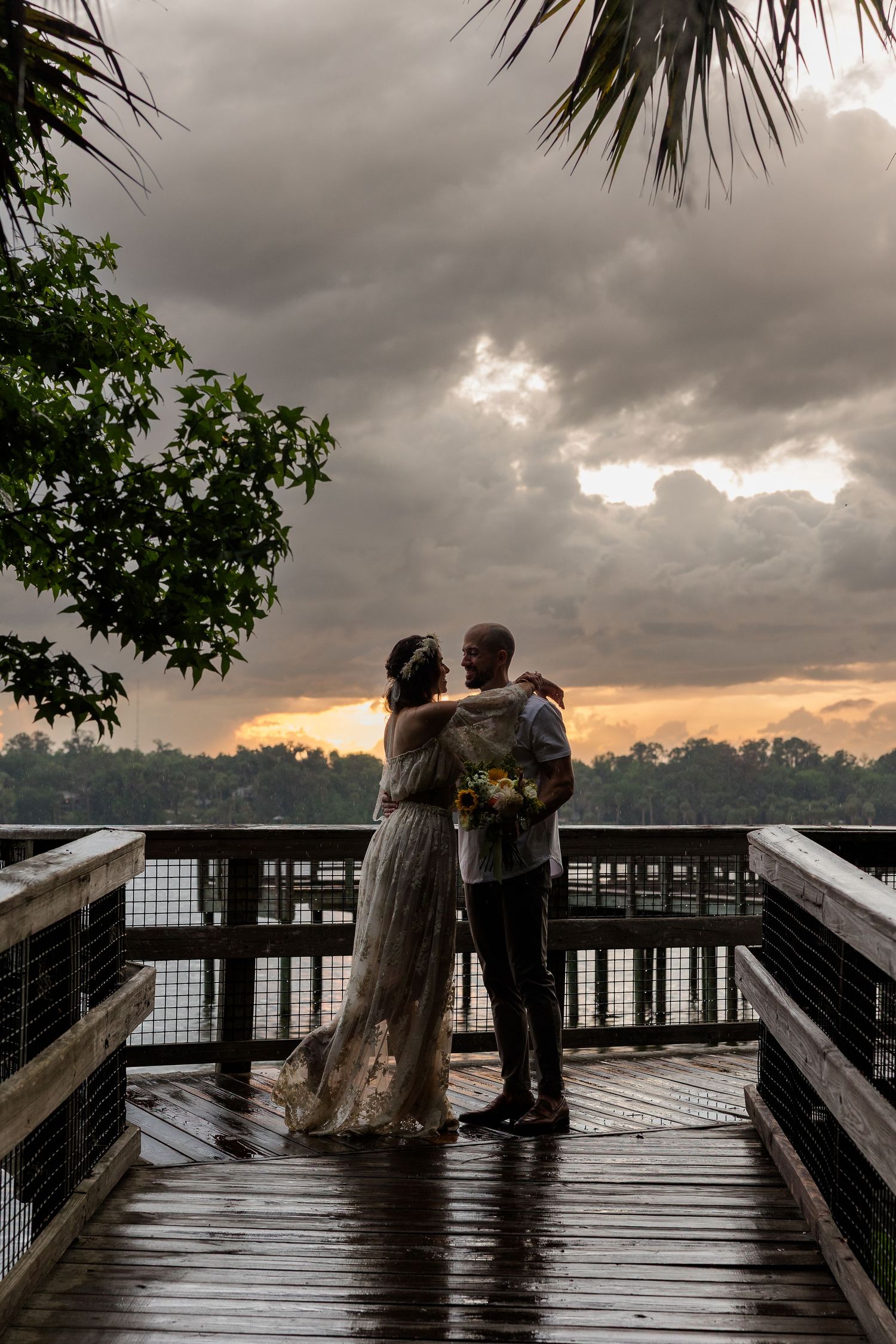 Couple embracing on a wet wooden dock at sunset with dramatic storm clouds over a lake, bride holding a bouquet.