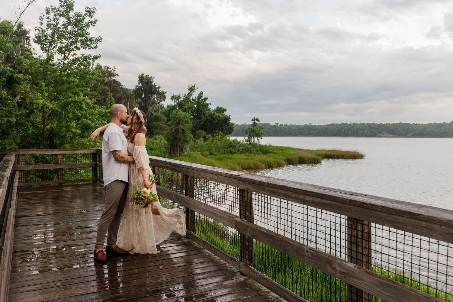 Couple sharing a romantic kiss on a wooden lakeside deck surrounded by lush greenery on a cloudy day.