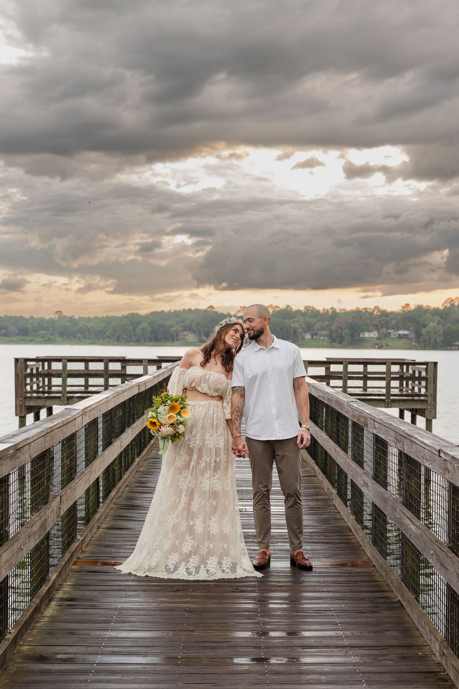 Bride and groom hold hands on a wooden pier by a lake under dramatic storm clouds at sunset.