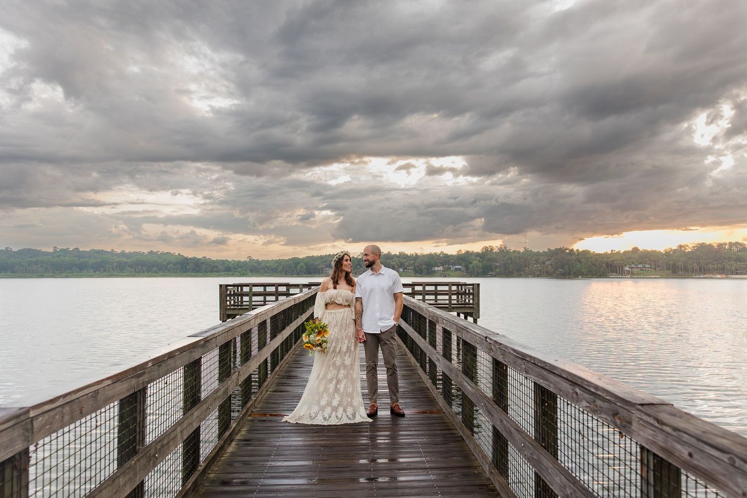 Bride and groom stand on a wooden pier overlooking a lake at sunset under dramatic cloudy skies.