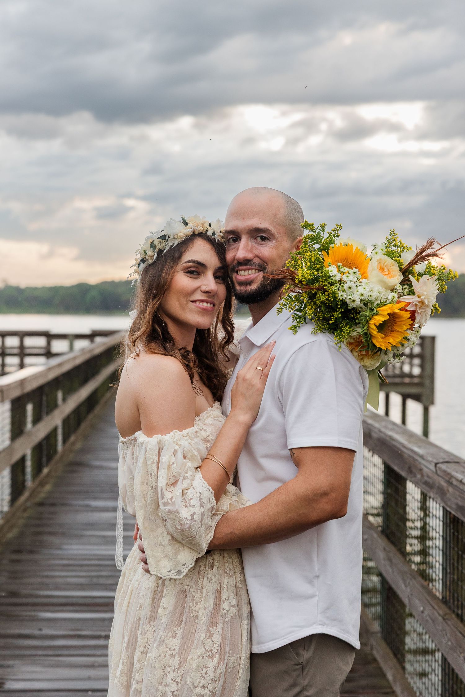 Bride and groom embracing on a wooden pier holding sunflower bouquet, bride wearing lace dress and floral crown.
