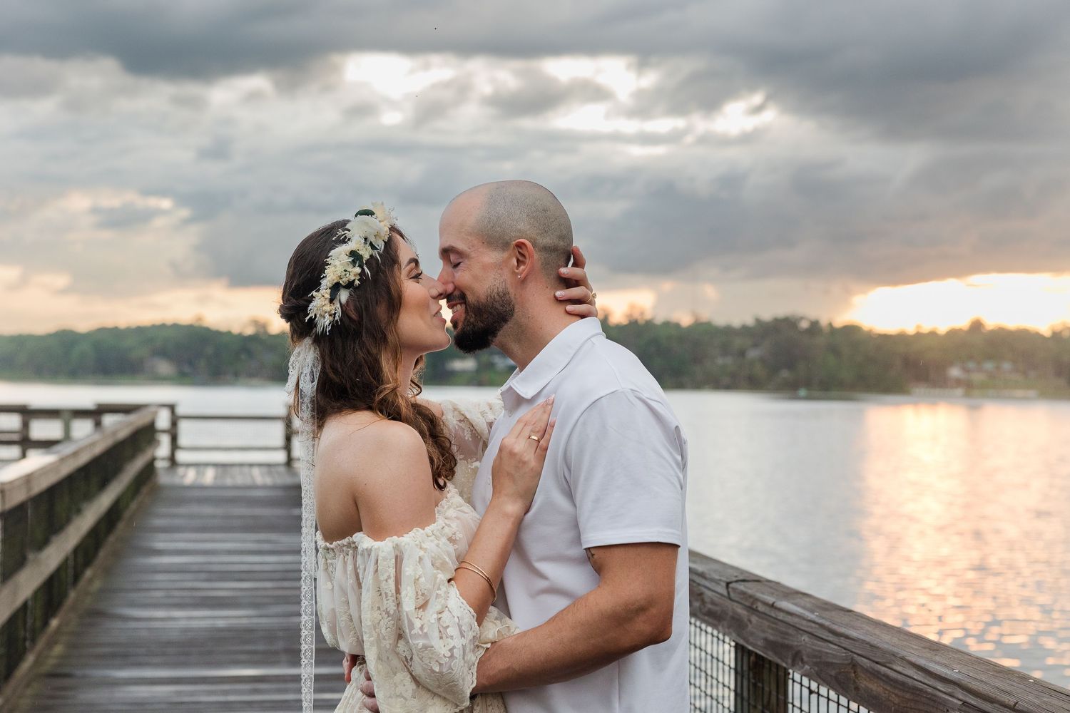 Couple sharing a romantic moment on a lakeside dock at sunset, bride wearing floral crown in outdoor engagement photo.