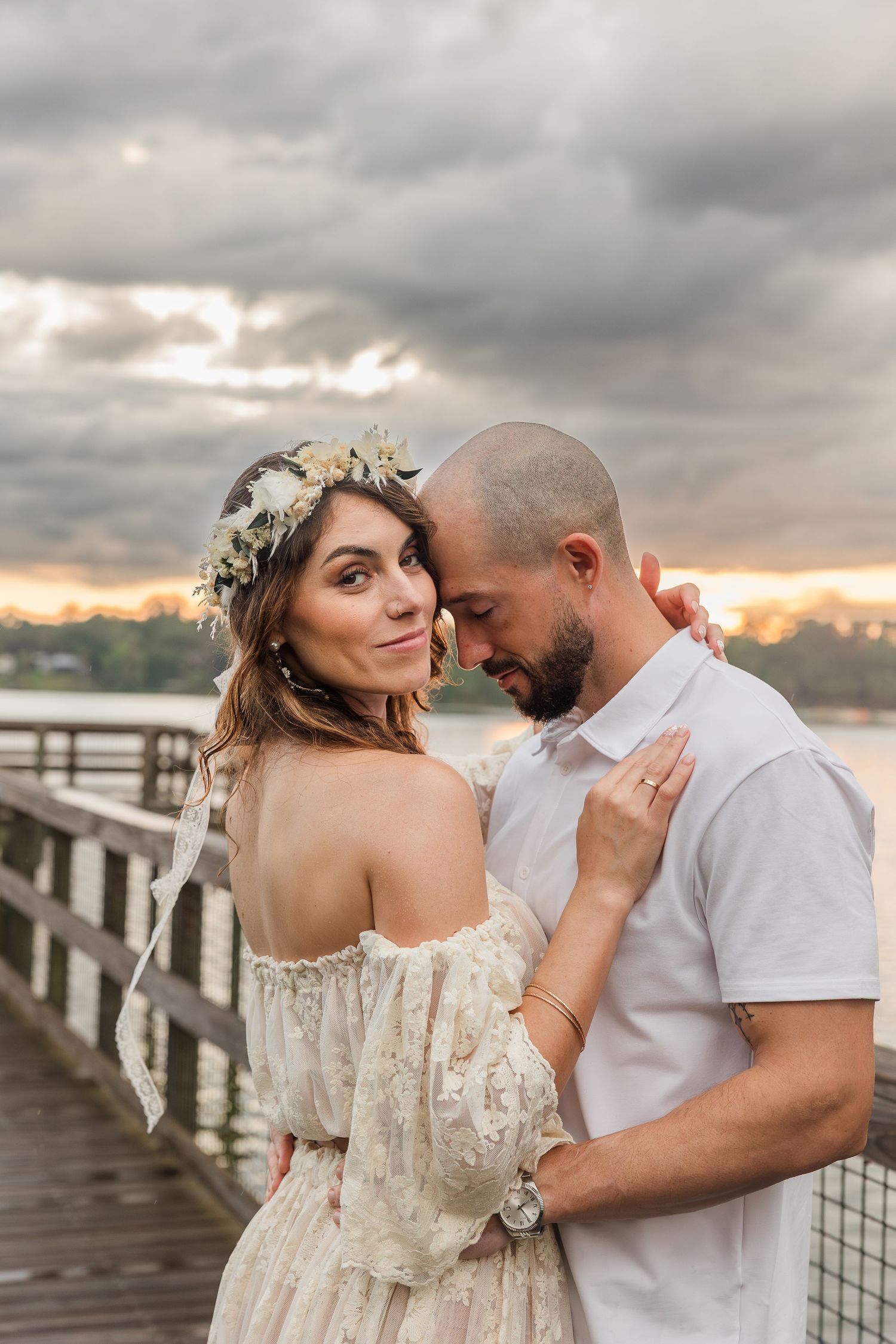 Couple embracing on a pier at sunset, woman wearing floral crown and off-shoulder dress, man in white shirt, cloudy sky.