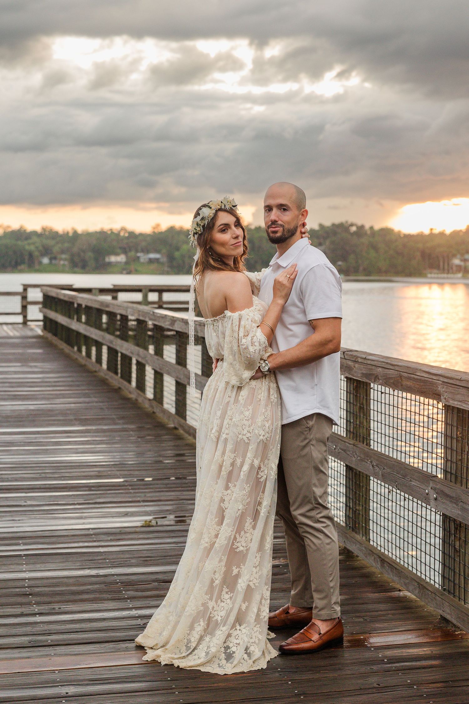 Bride and groom embrace on a wooden pier at sunset, bride wearing lace gown and floral crown.