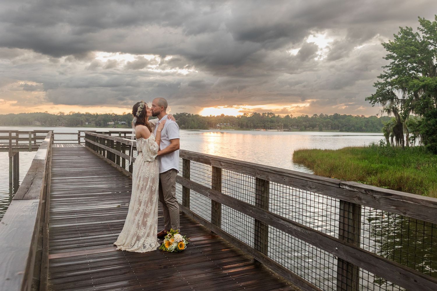 Couple shares a kiss on a wooden dock at sunset with a scenic lake and dramatic cloudy sky backdrop.