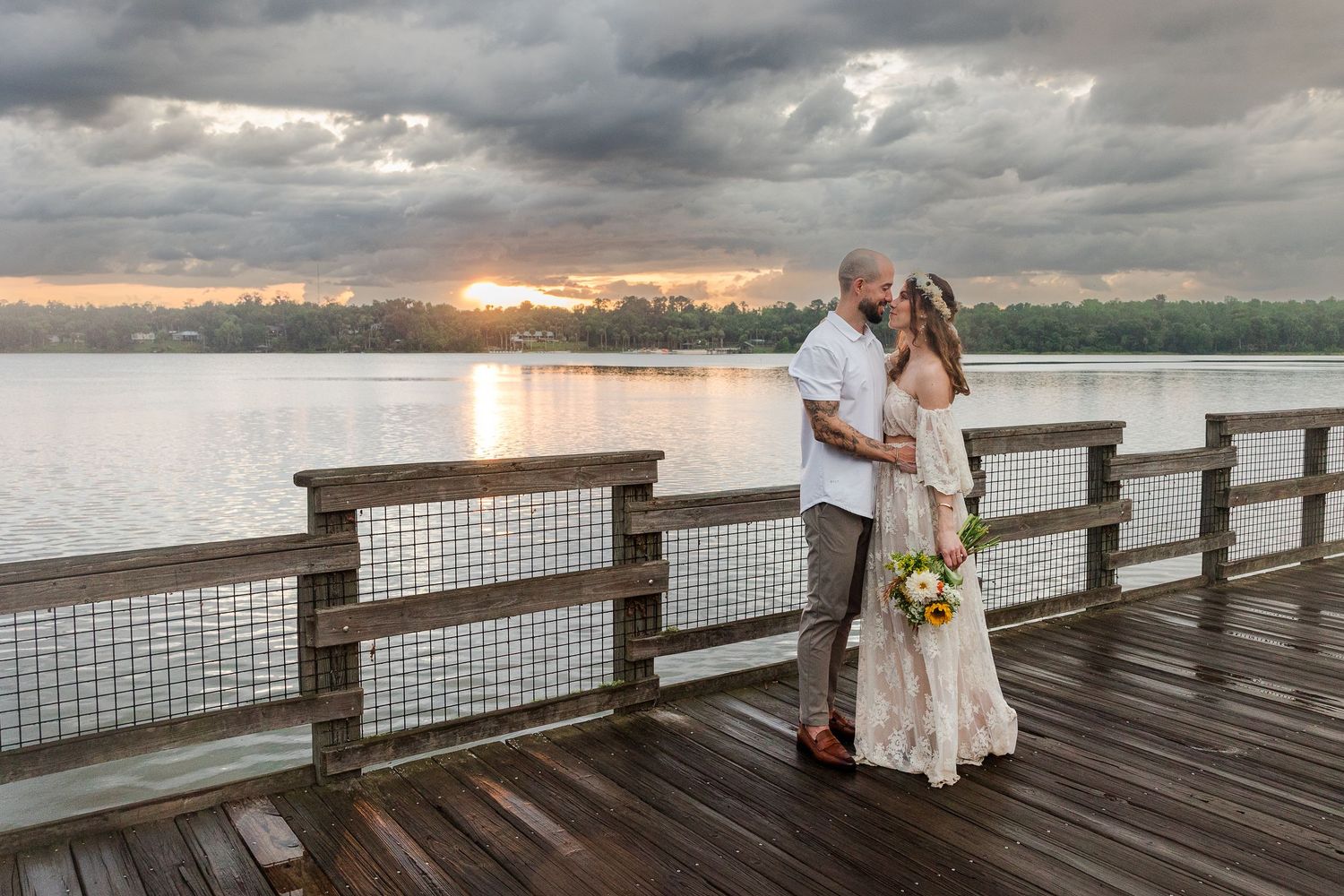 Couple sharing a romantic kiss on a waterfront dock at sunset, bride holding a bouquet in a lace gown.